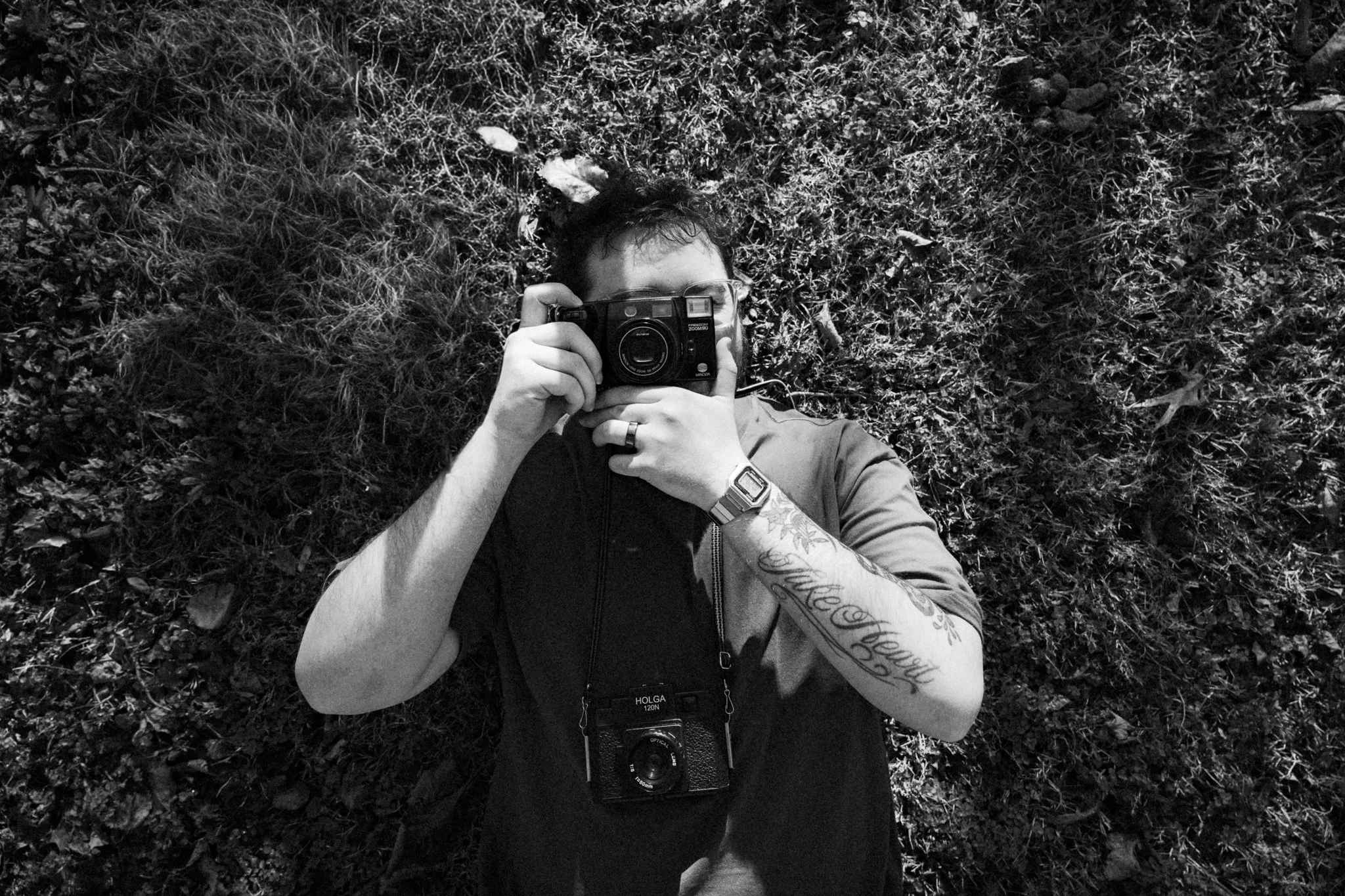 A person lying on the ground, taking a photograph with a camera, surrounded by grass and foliage.