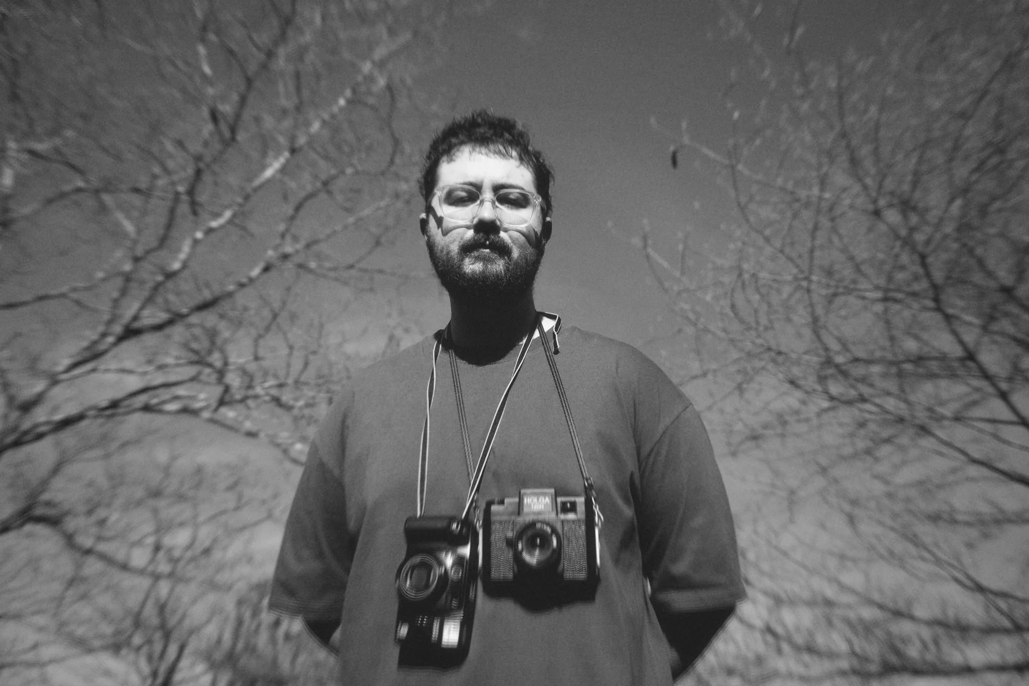 A man with glasses and a beard stands outdoors during daytime, wearing a t-shirt and a camera hanging around his neck, with leafless trees in the background.