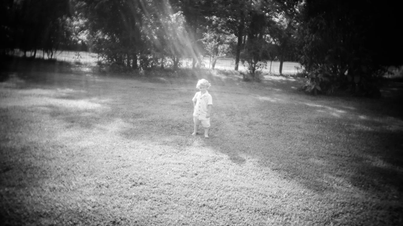 A young child standing on a grassy lawn with trees and a fence in the background, sunlight streaming through the trees, in black and white.