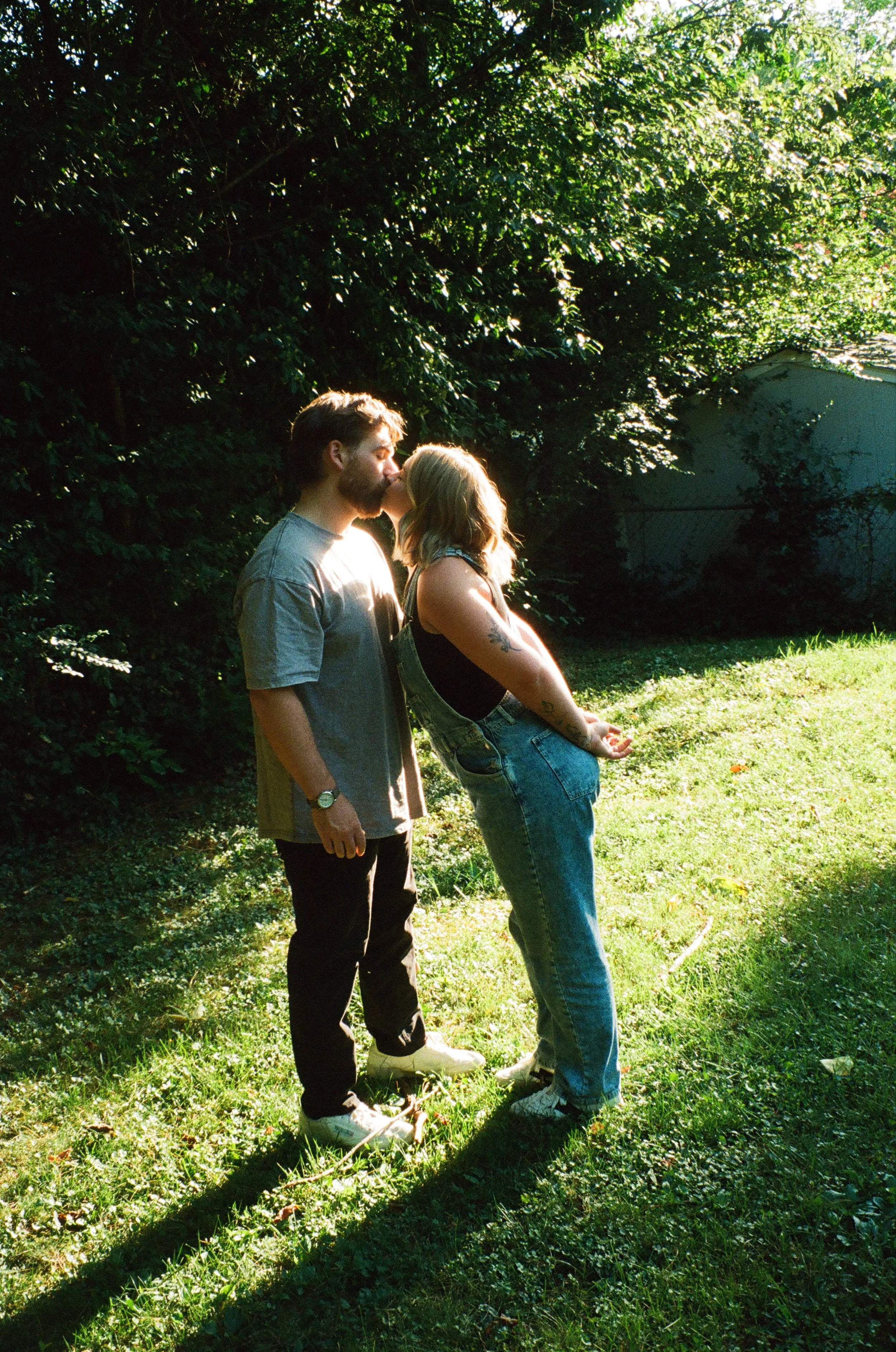 A man and woman are kissing on a grassy lawn in sunlight, with trees and a shed in the background.