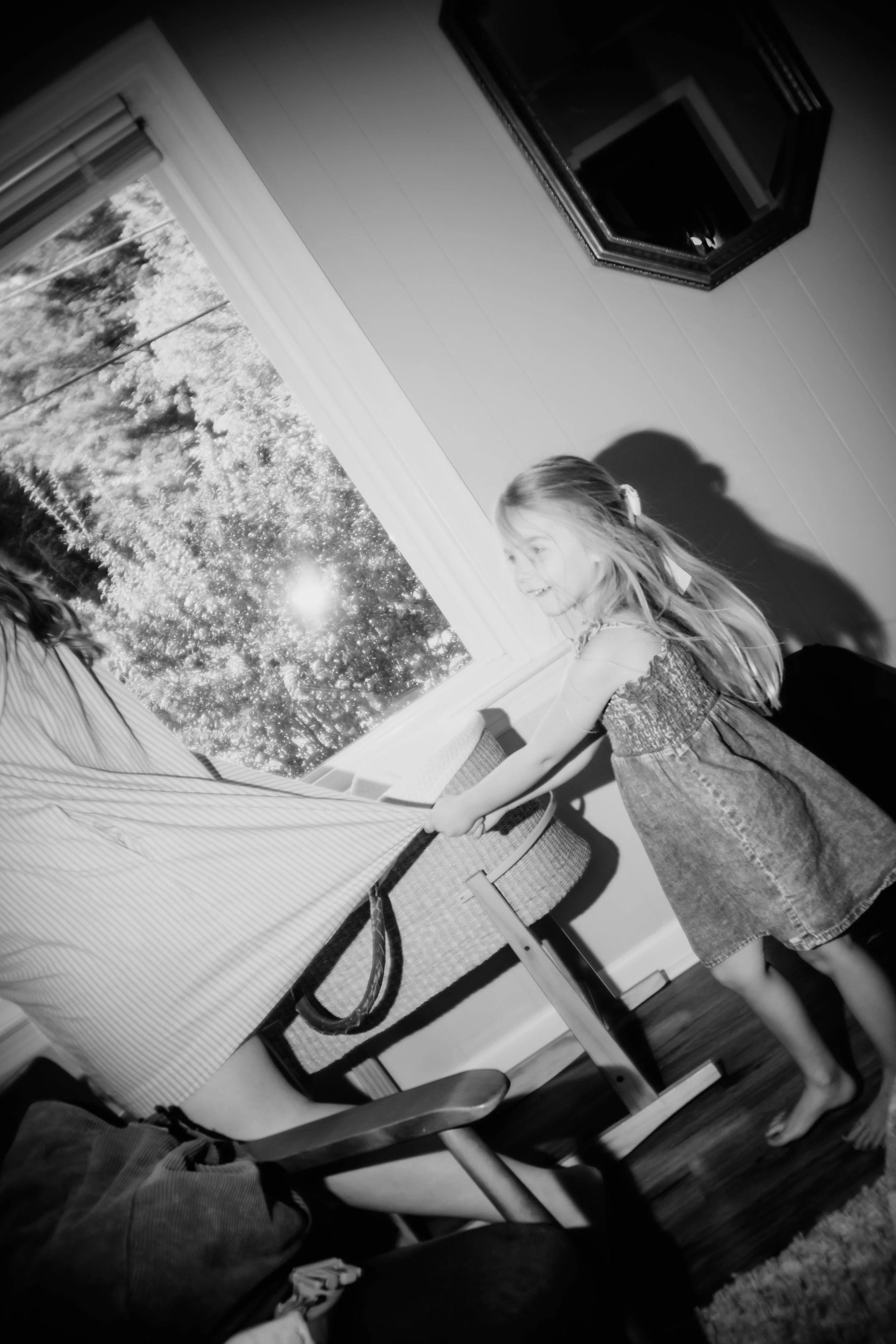 A young girl with blonde hair tied back, wearing a dress, is pulling a tablecloth off a table near a window with trees outside. The room has a wall-mounted television and a mirror on the wall.