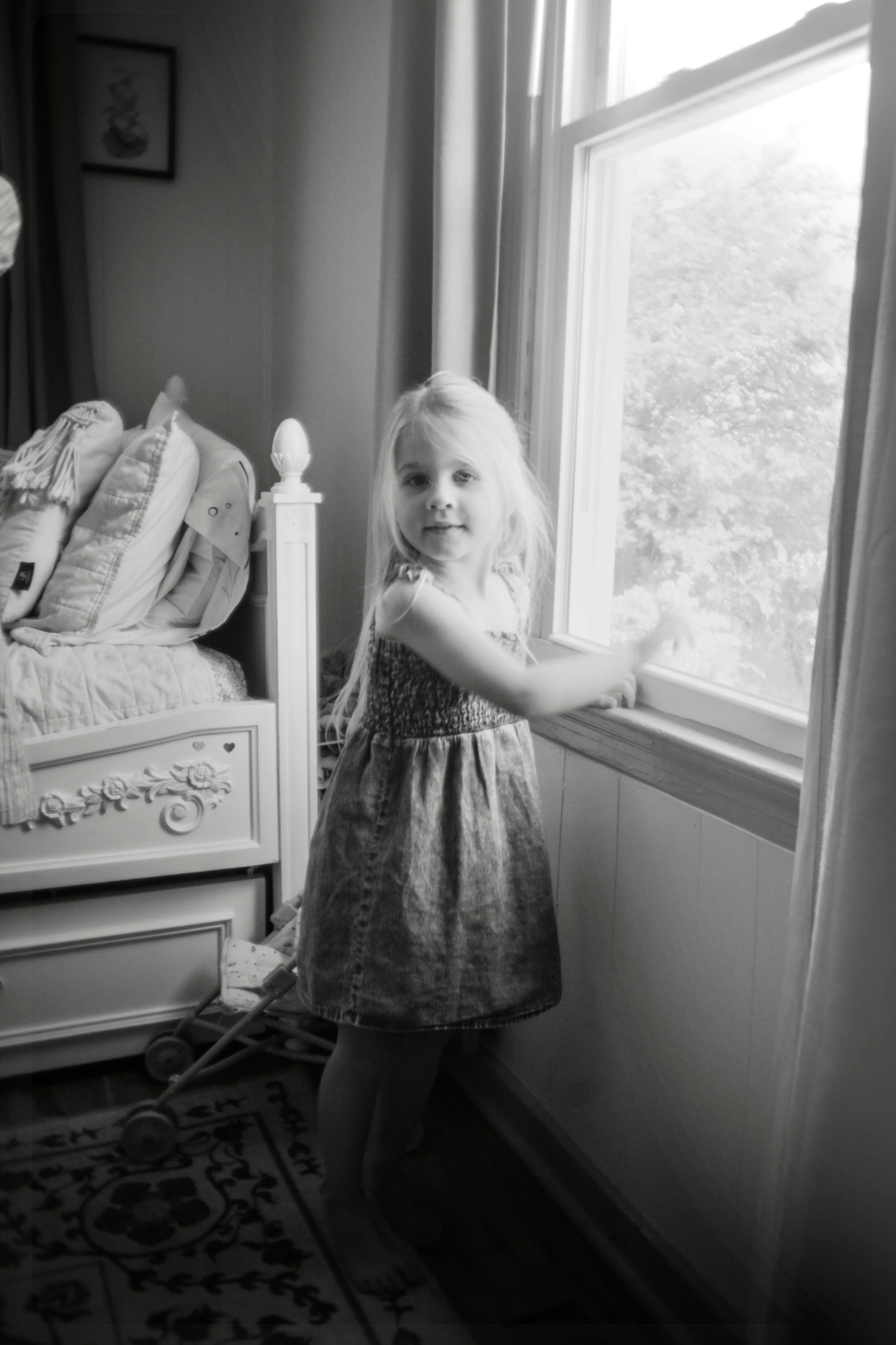 A young girl with long light-colored hair standing by a window in a bedroom, looking at the camera. The room has a bed with pillows, a framed picture on the wall, and a small stroller on the floor.