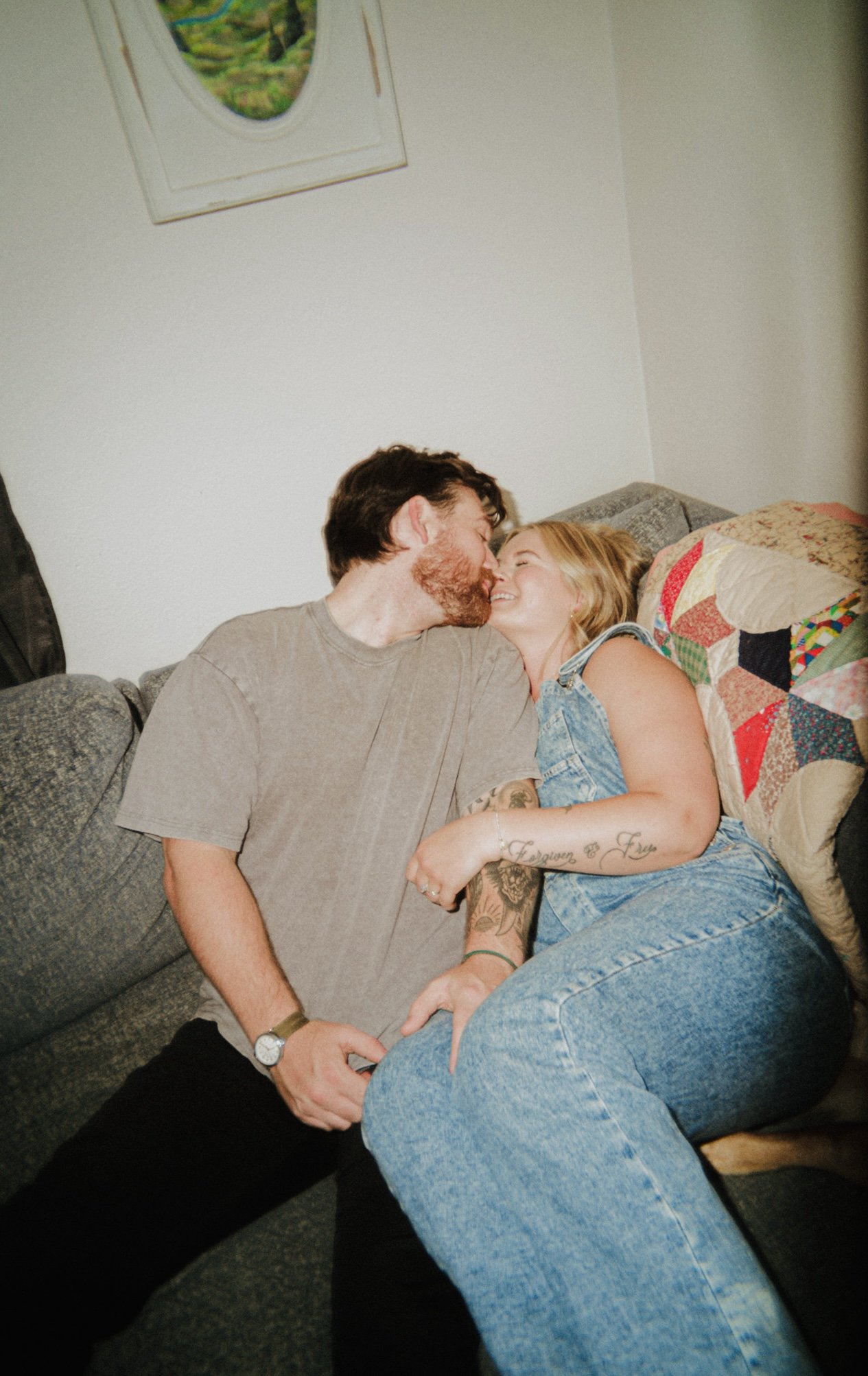 A couple sitting on a gray couch, leaning in for a kiss, in a cozy living room.
