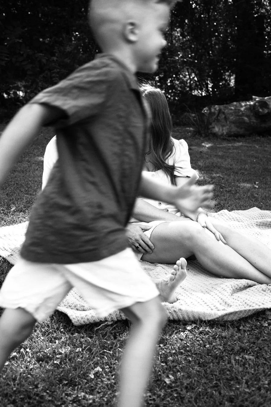 A boy is running in front of a woman sitting on a blanket outdoors. The woman, with long hair, is sitting with her hands on her knee. The scene is in a natural setting with trees in the background, and it is captured in black and white.