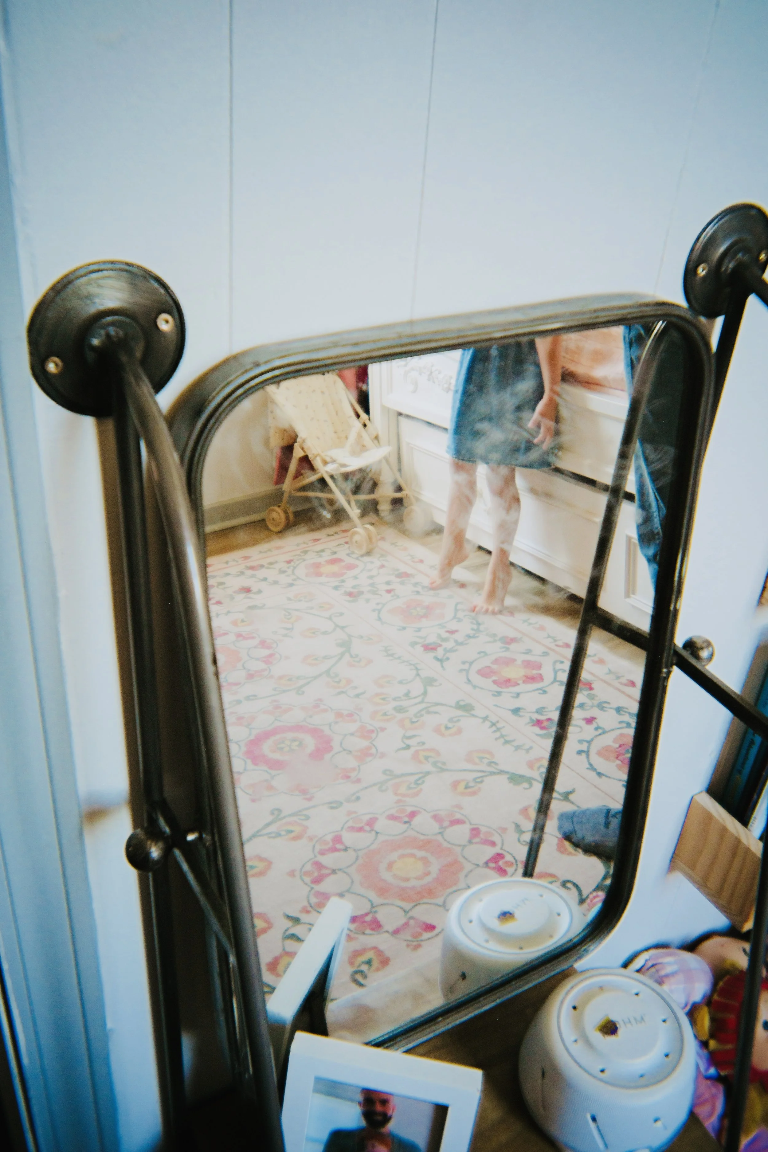 Reflective surface of a standing mirror showing a person in a pink shirt and denim shorts holding a phone, with indoor furniture including a doll stroller and a colorful patterned rug.