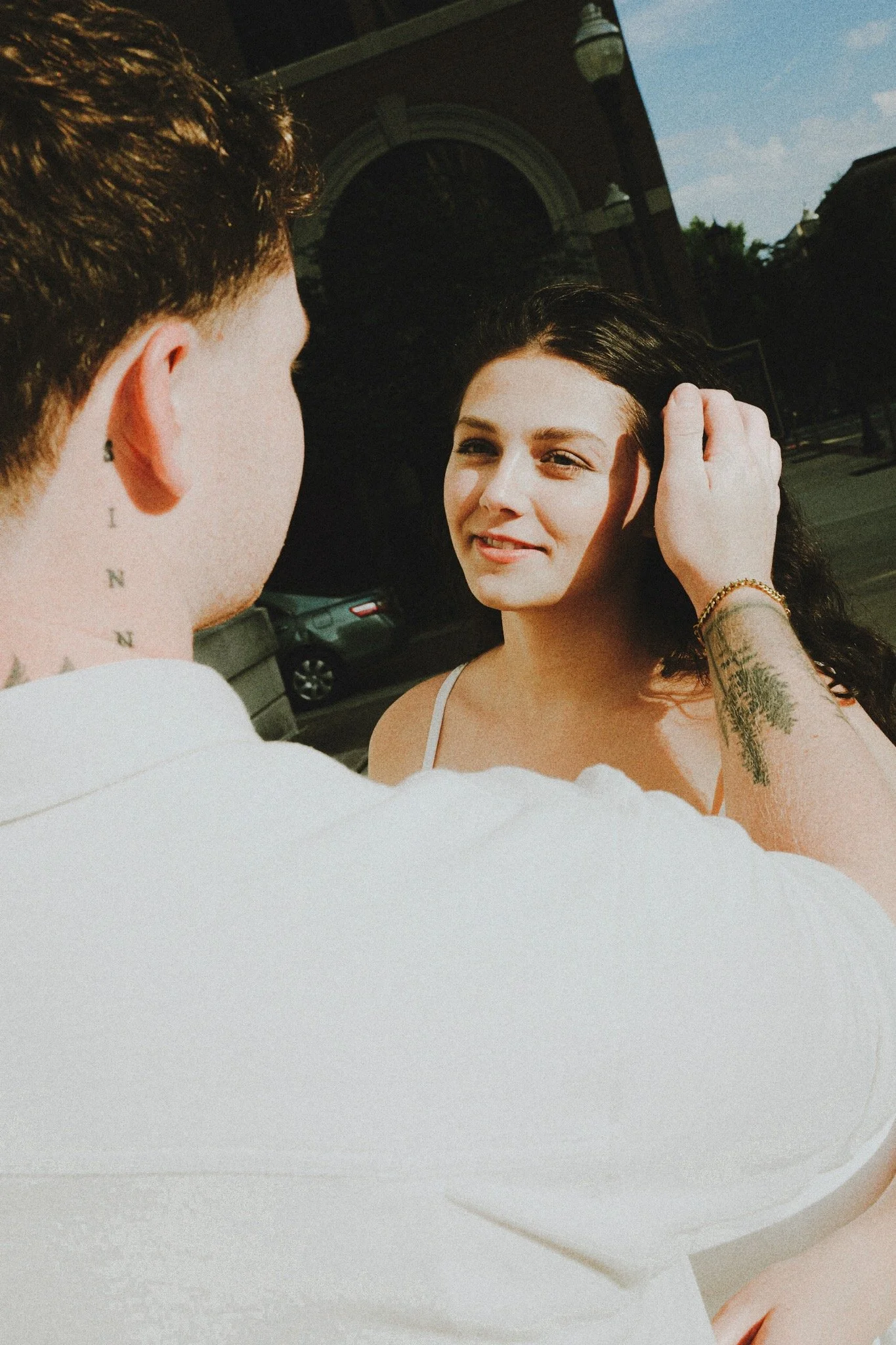A man is touching the hair of a woman with dark curly hair, standing outdoors in sunlight, with a building and a car visible in the background.