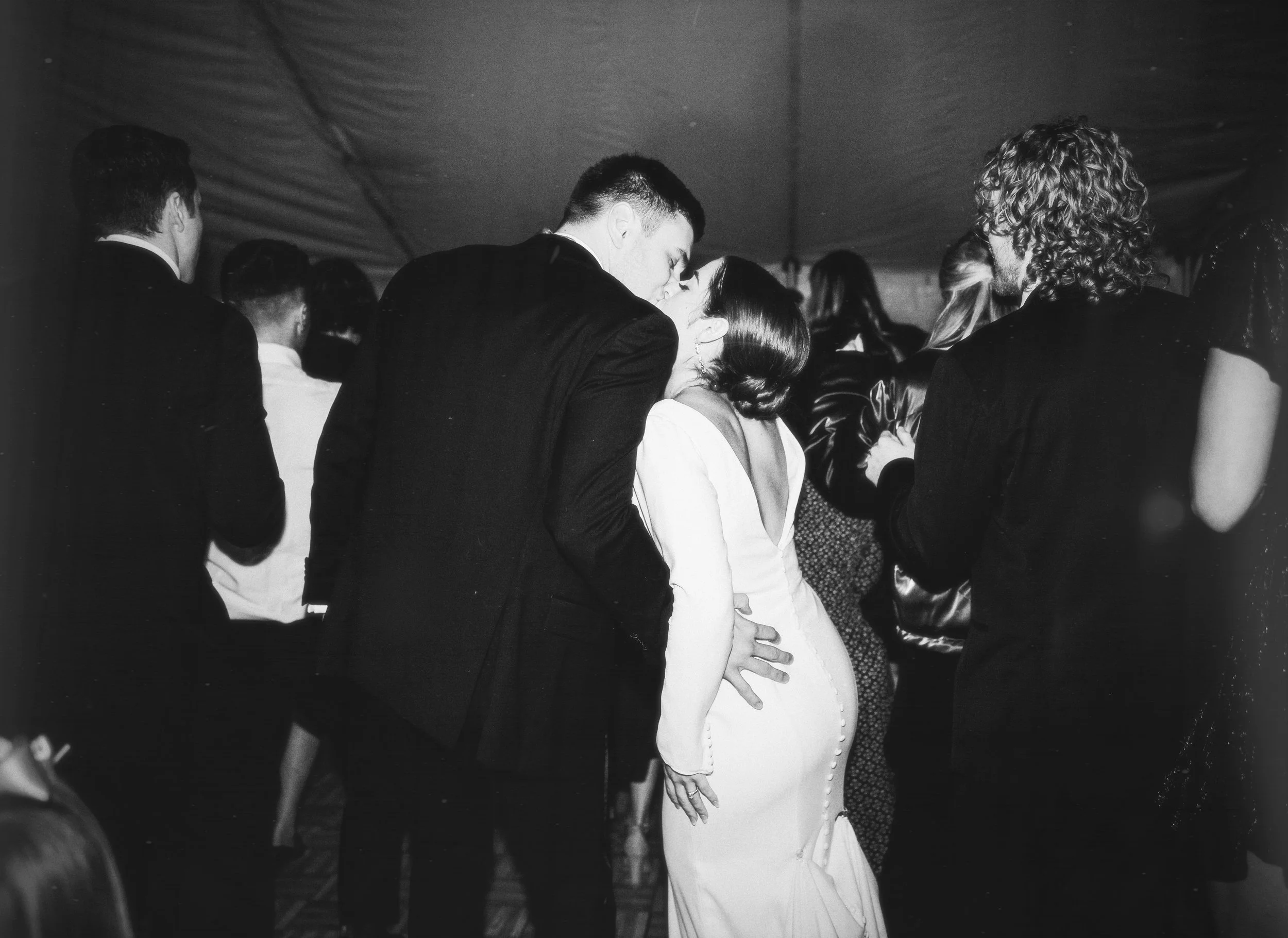 A black-and-white photo of a couple kissing at a formal event, surrounded by other people in tuxedos and dresses.