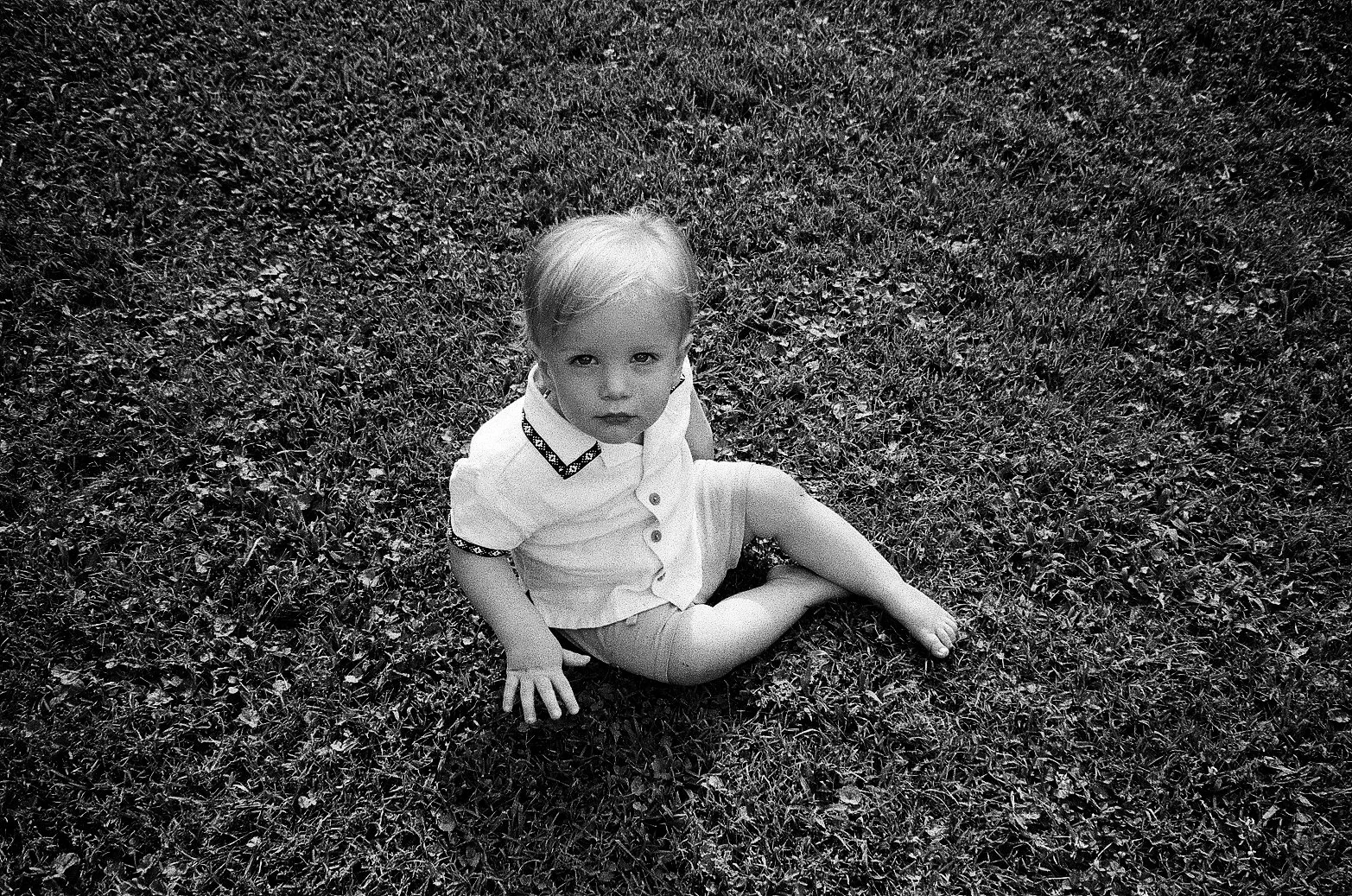 A black and white photo of a young child sitting on grass, looking up at the camera with a serious expression.