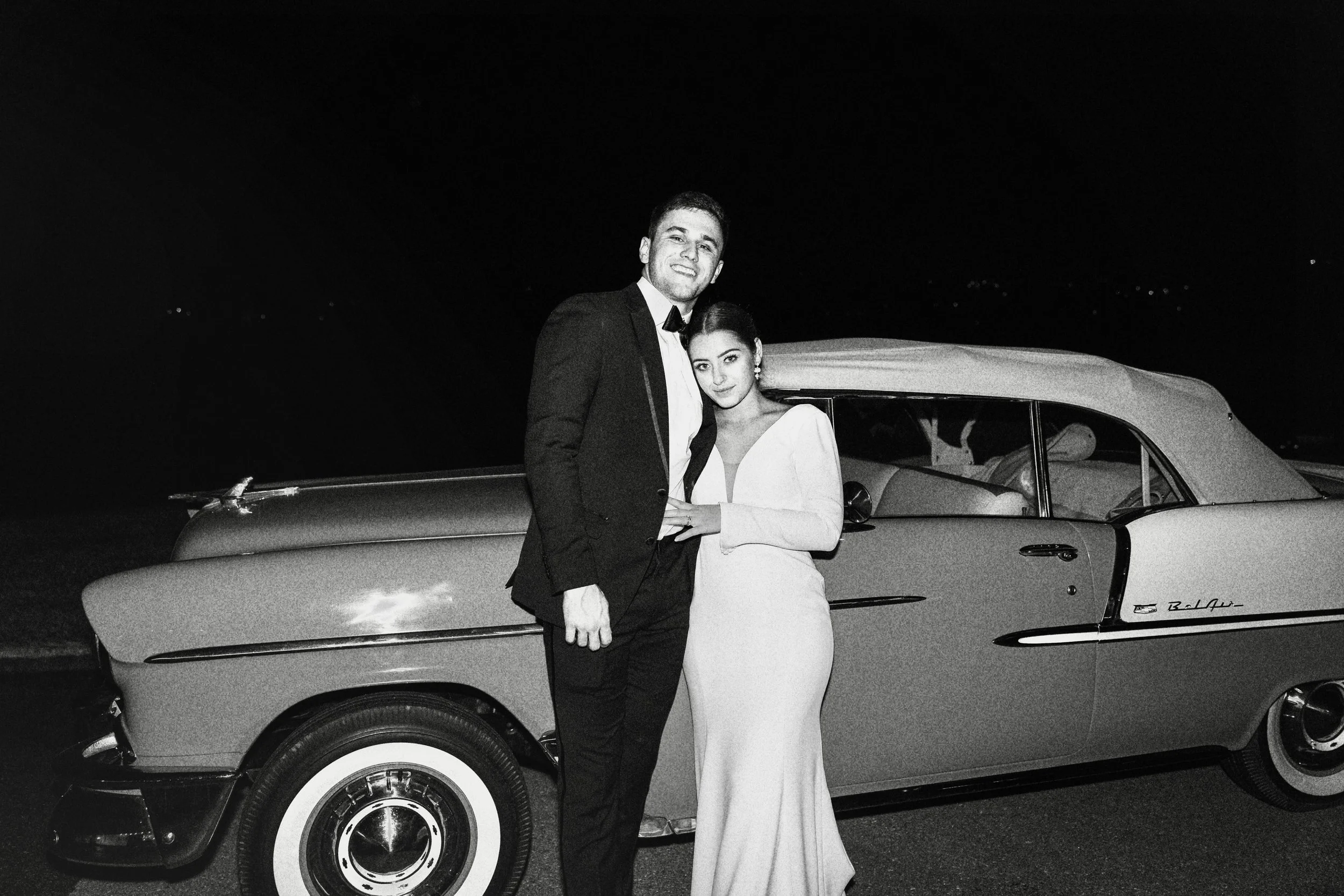 A man in a tuxedo and a woman in a formal white gown standing together at night in front of a classic car.