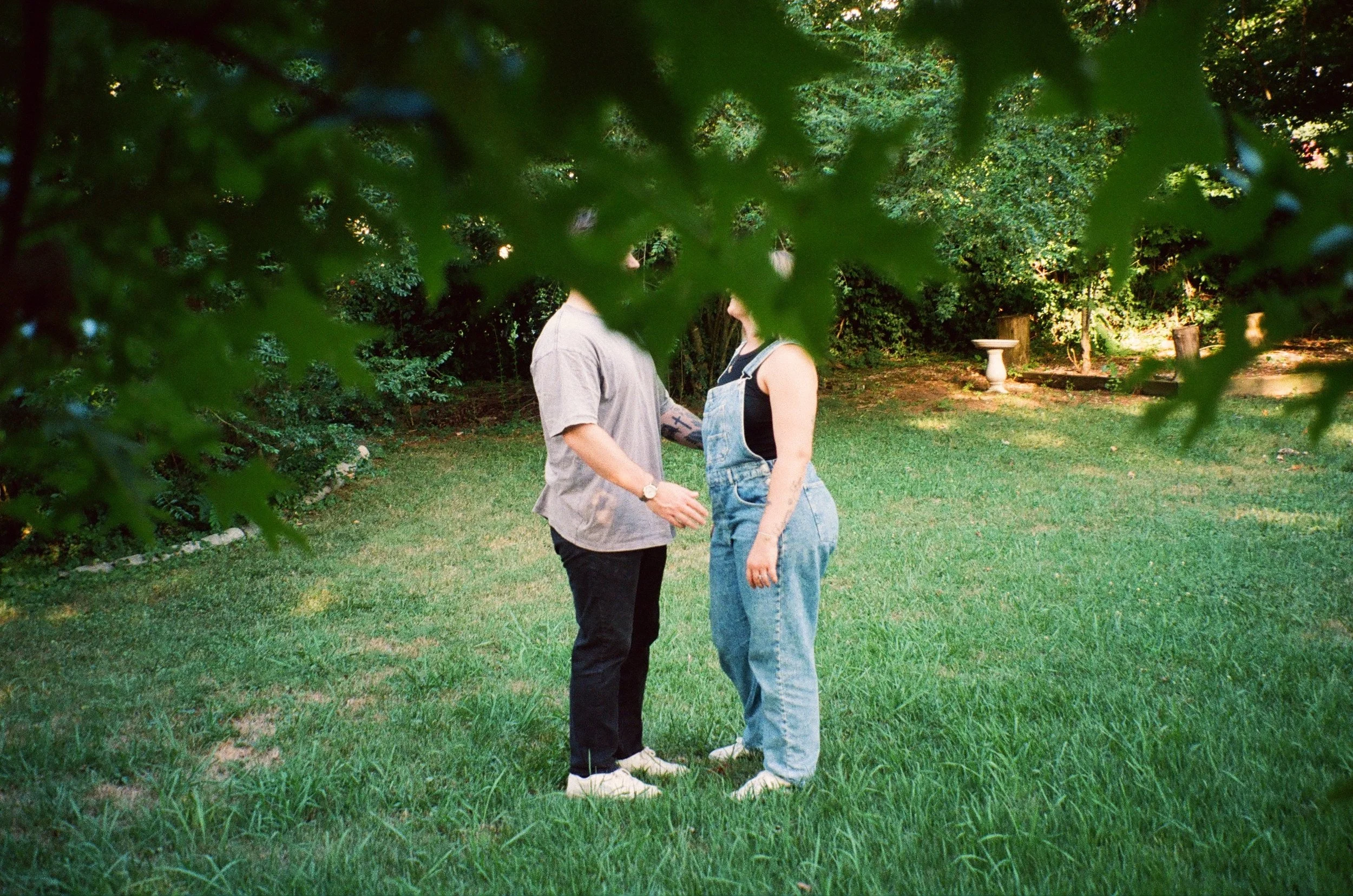 Two people standing close together on a grassy lawn, partially obscured by green leaves in the foreground, with trees and garden ornaments in the background.