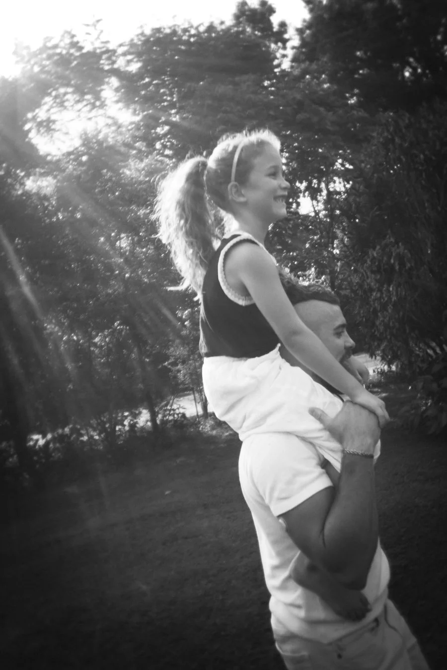 A man is giving a girl a piggyback ride in a park, and they are both smiling. The sun is shining through the trees.
