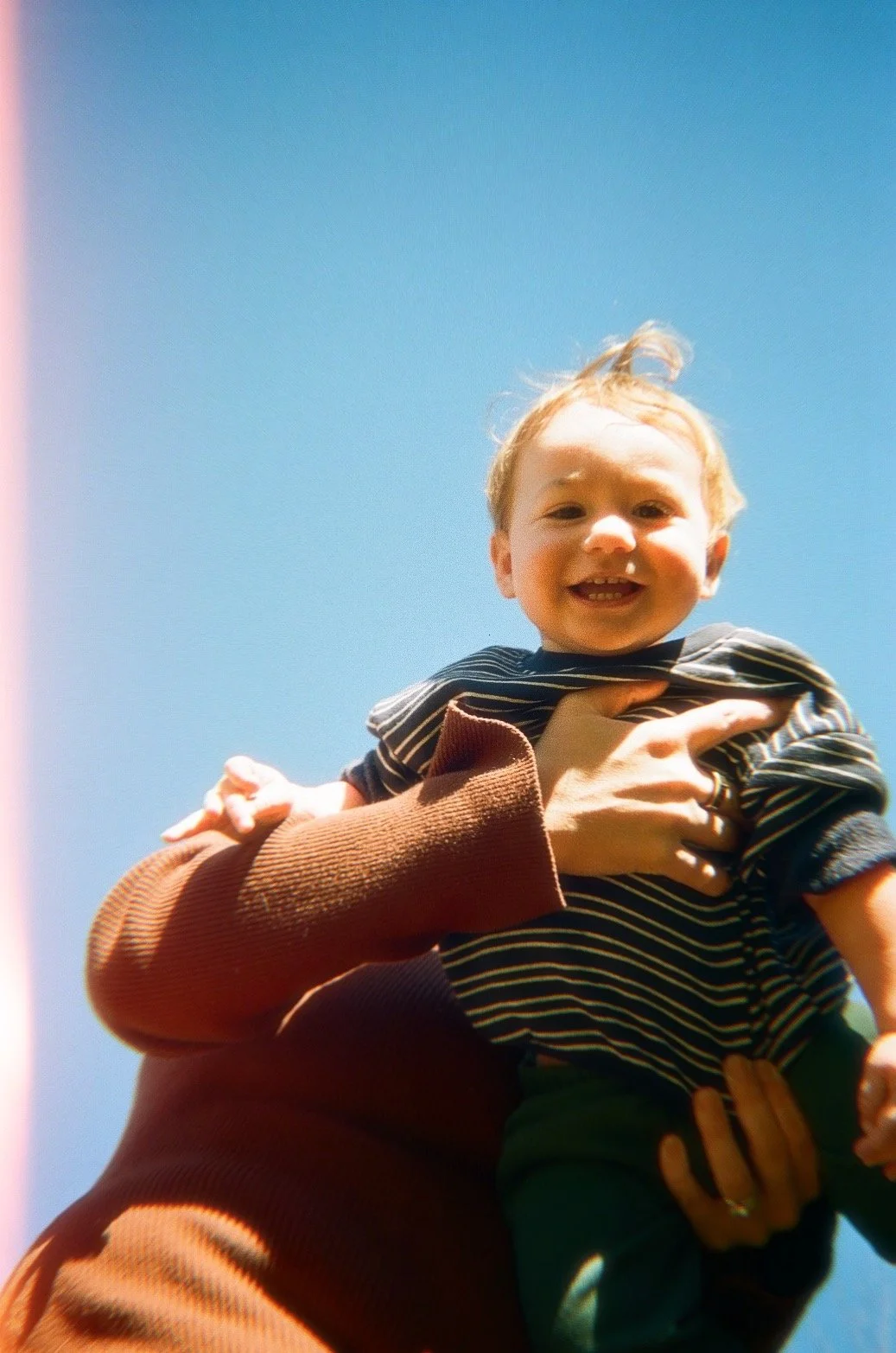 A smiling toddler being lifted into the air against a clear blue sky.
