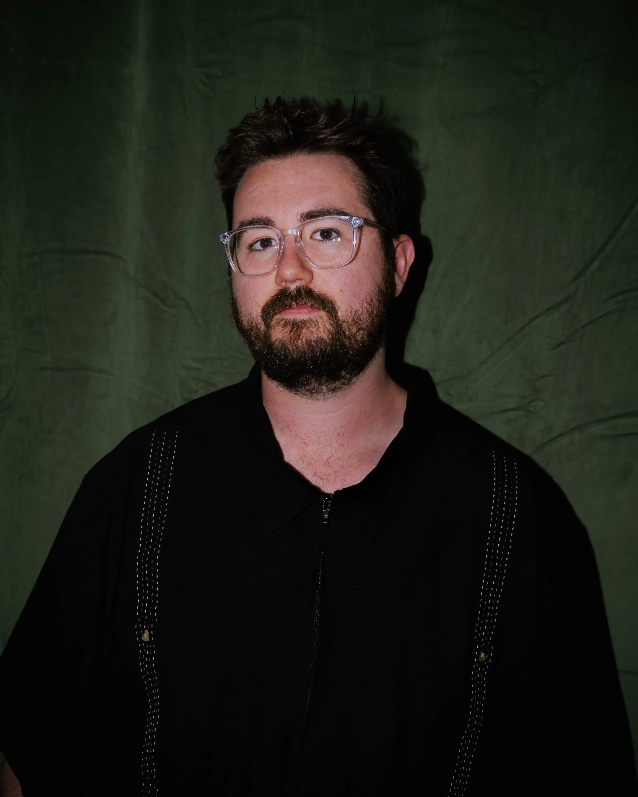 Portrait of a bearded man with glasses, dark hair, wearing a black shirt, against a dark green background.