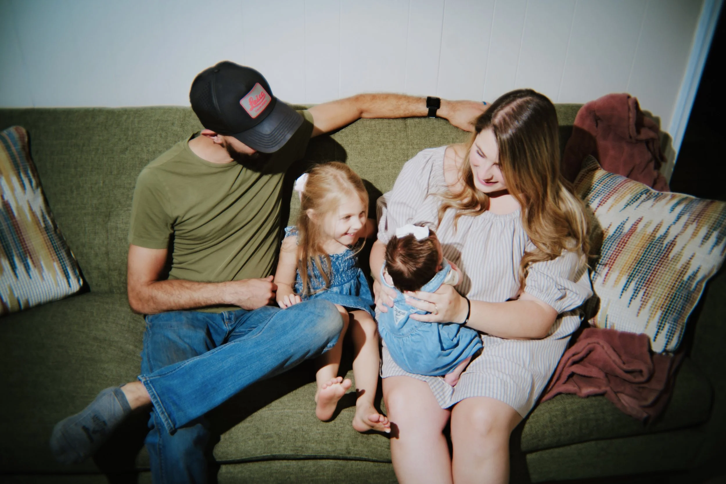 A family of four sitting on a green couch, with a man wearing a black cap, a young girl, a woman holding a baby, and two patterned pillows.