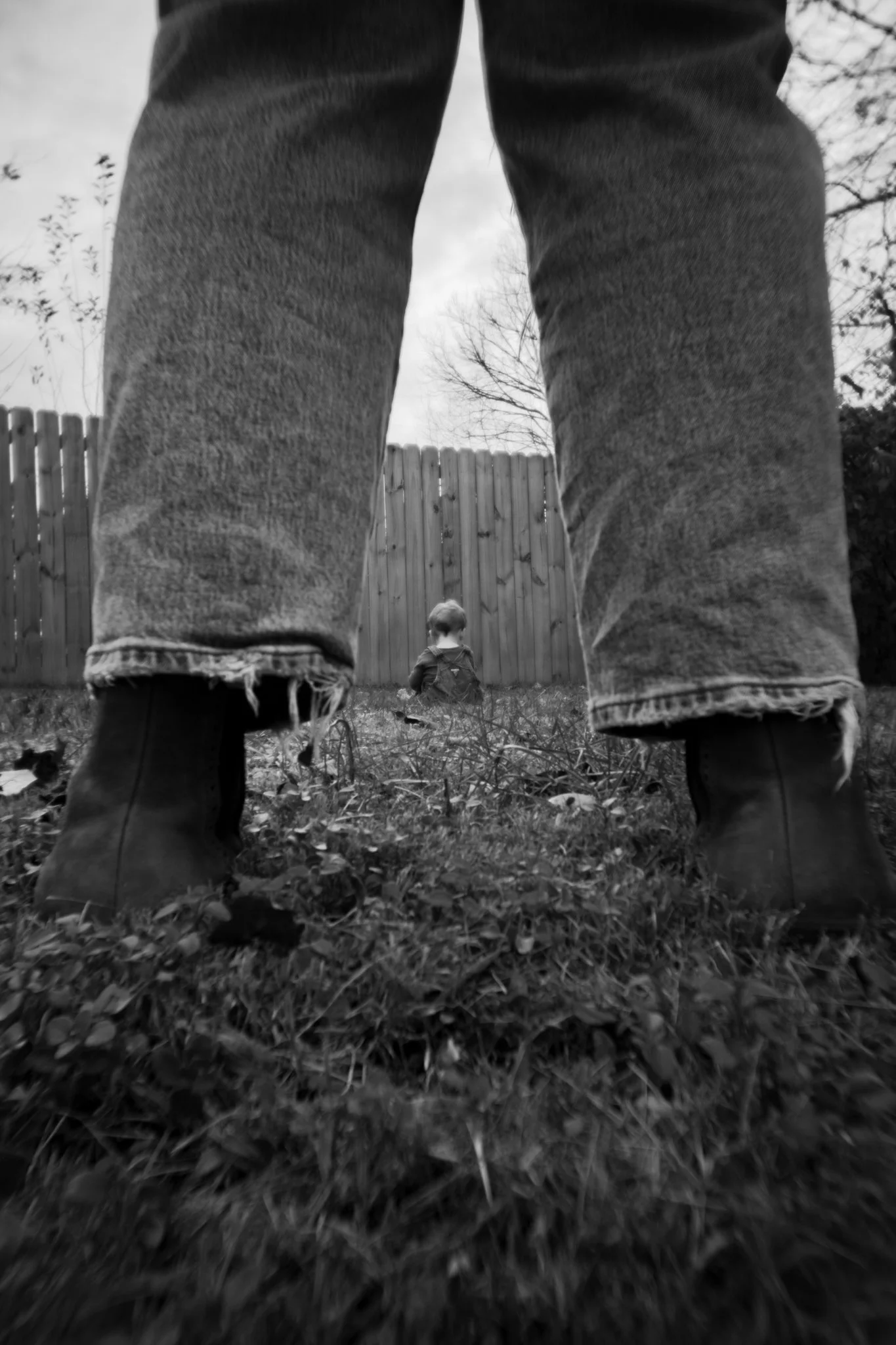 Black and white photo showing a small child sitting on the grass in a backyard, framed by large legs and boots of an adult standing nearby, with a wooden fence and trees in the background.