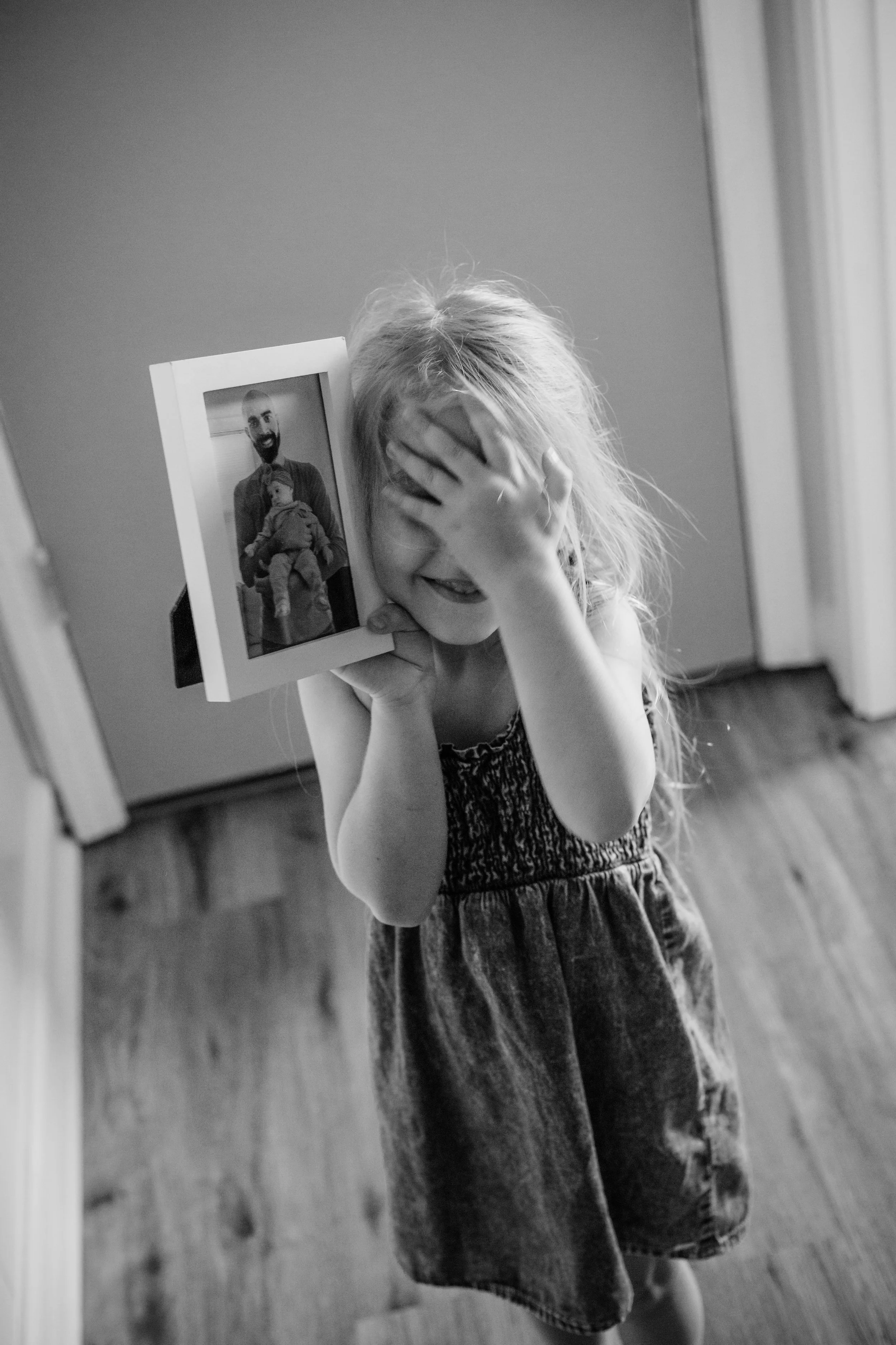 A young girl with long hair is smiling and covering her face with one hand while holding a picture of a man holding a child.