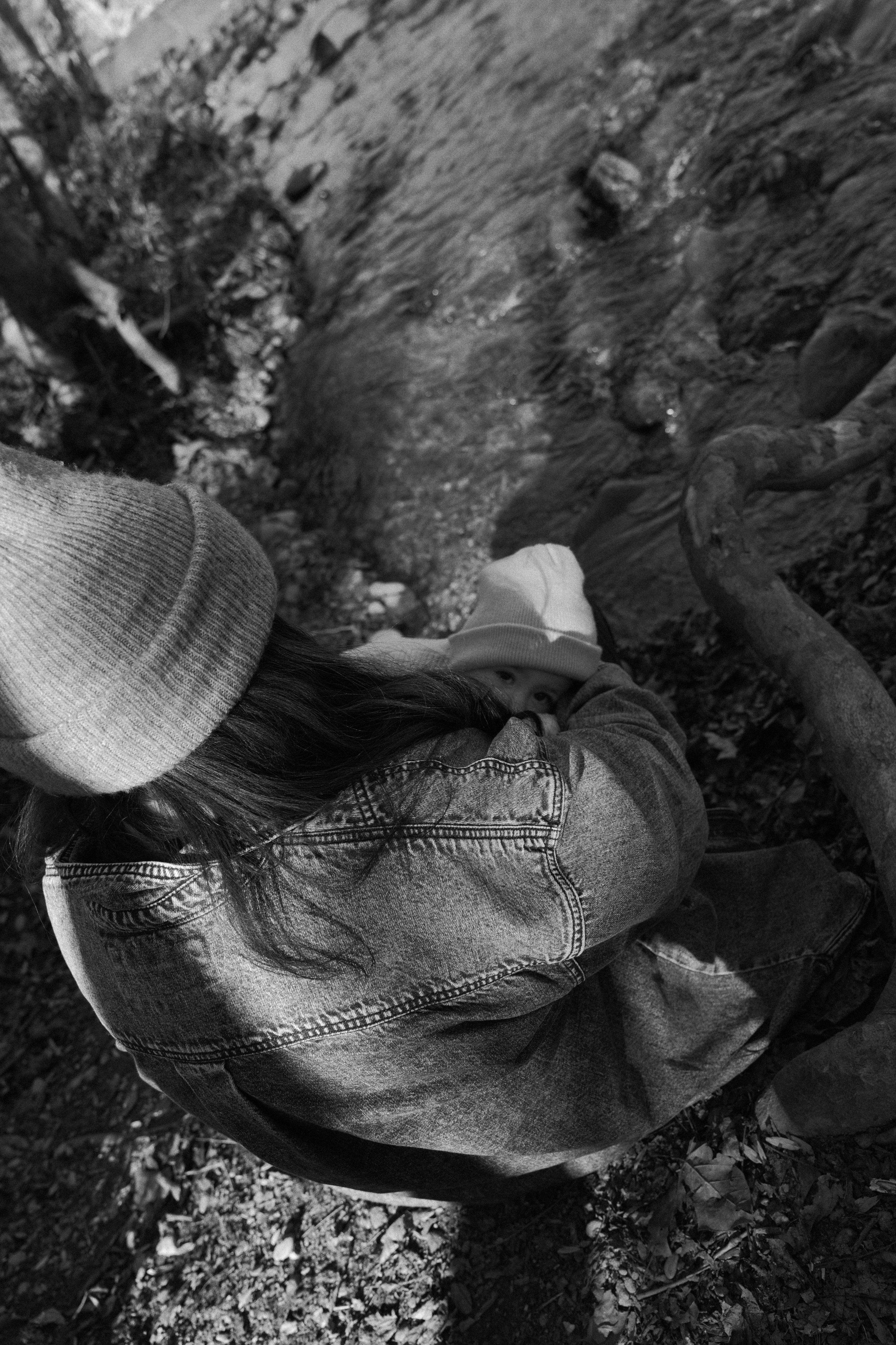 A black and white photo of a person with long hair wearing a denim jacket and a beanie, sitting beside a small stream or creek in a wooded area, with a child partially visible, wearing a light-colored hat, peering over their shoulder.
