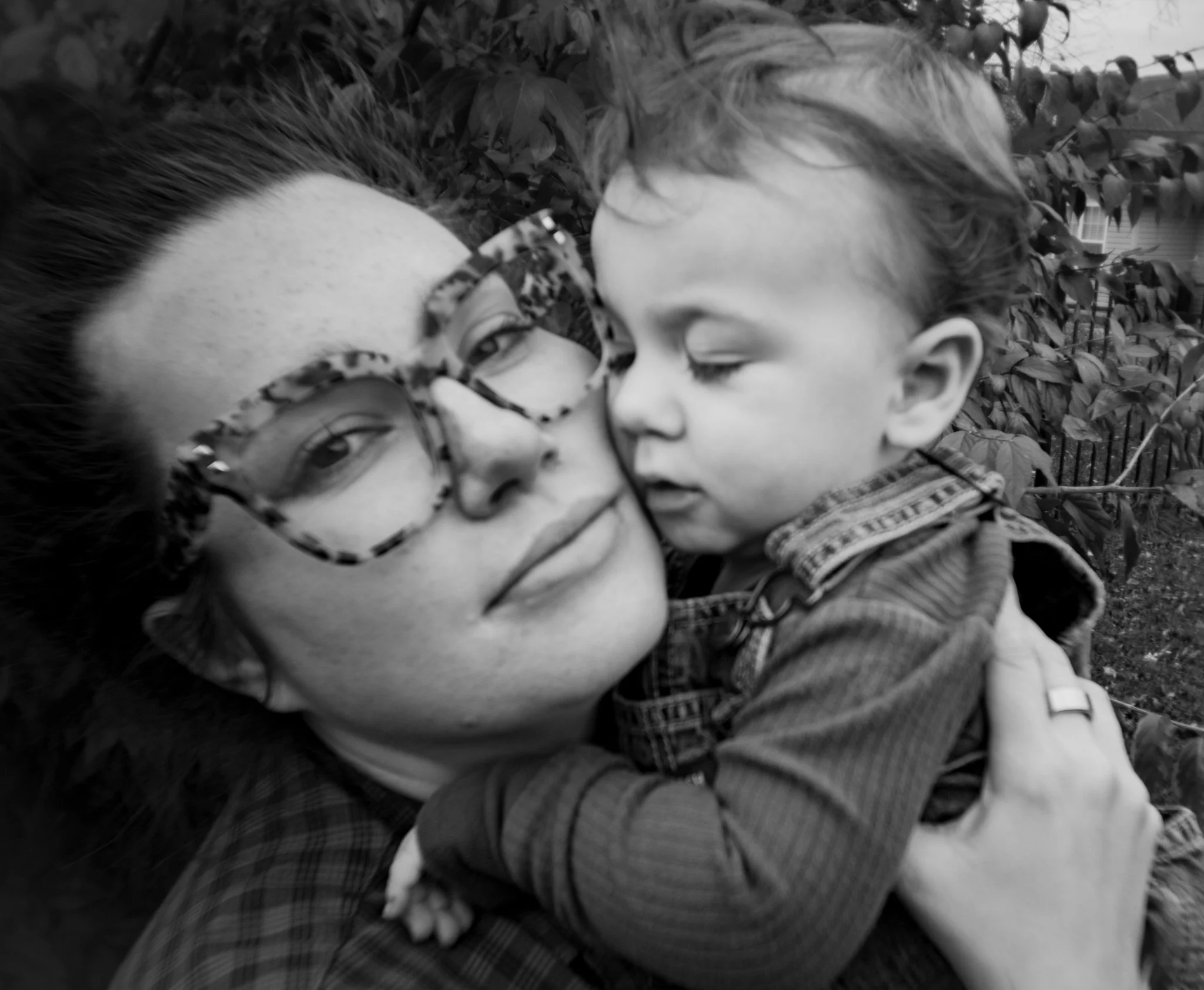 A woman wearing glasses and a kid hugging her, both with gray hair, outdoors, with trees and a fence in the background.