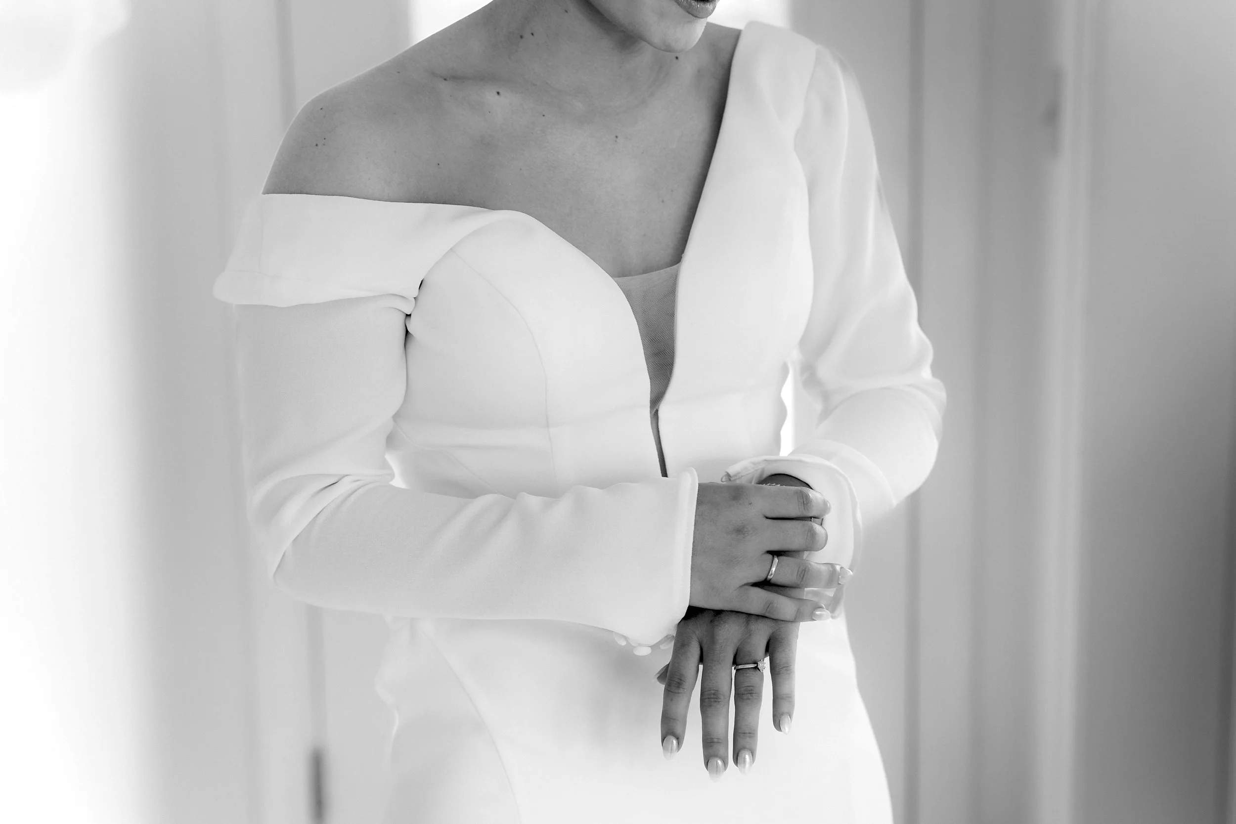 Black and white photo of a woman in an off-shoulder white dress, with her hands clasped in front of her, standing indoors.
