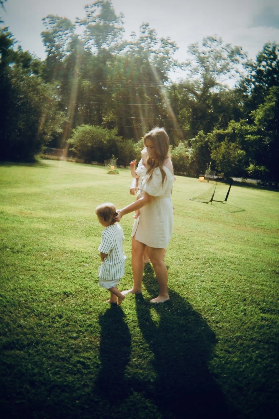 A woman and two children outdoors on a sunny day, standing on grass with trees in the background. The woman is holding one child's hand while the other child stands nearby, all barefoot.