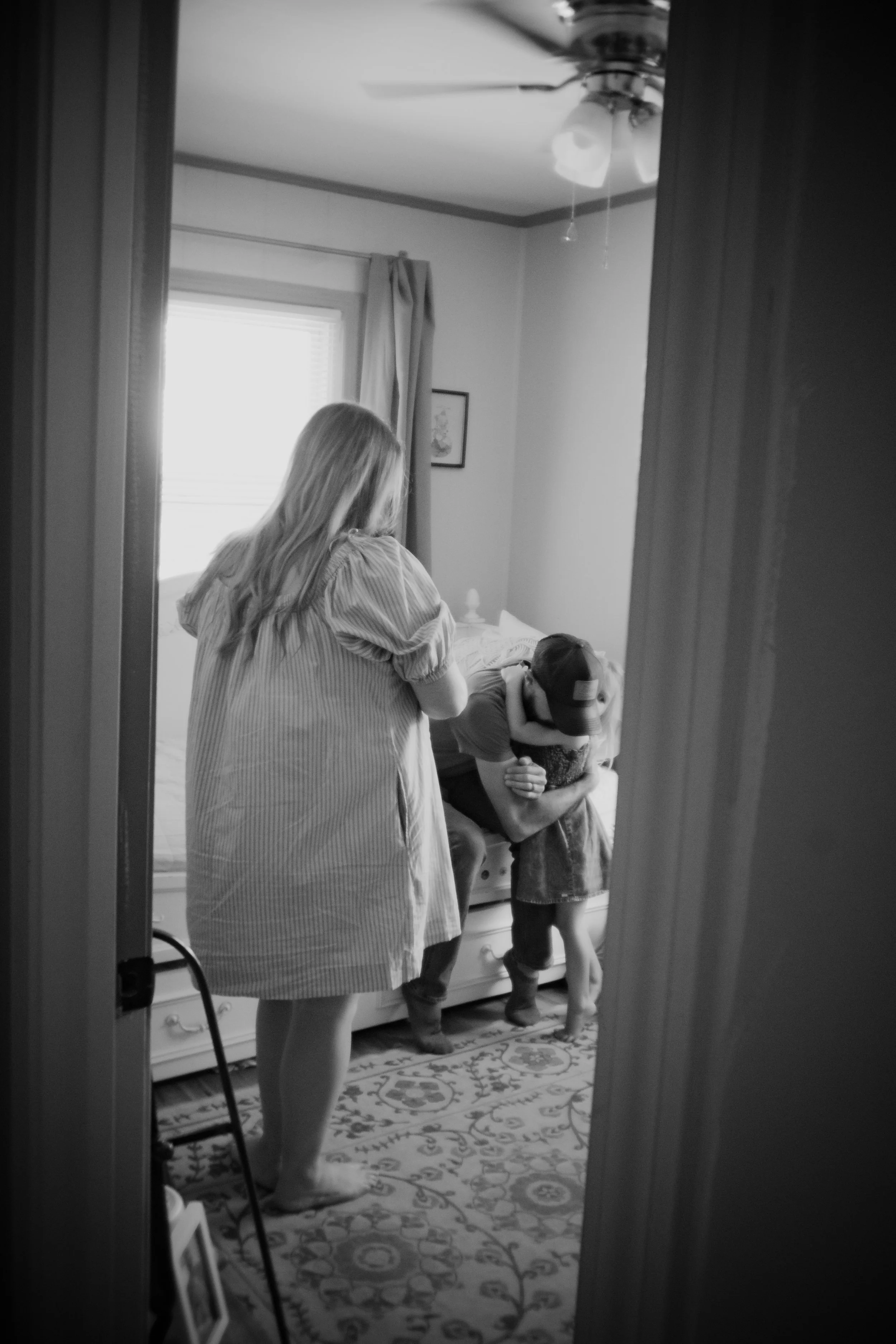 A woman and a child hugging each other in a bedroom, viewed through a doorway. The woman is standing and the child is hugging her with a hat on. There is a bed, a window, and framed art on the wall in the background.