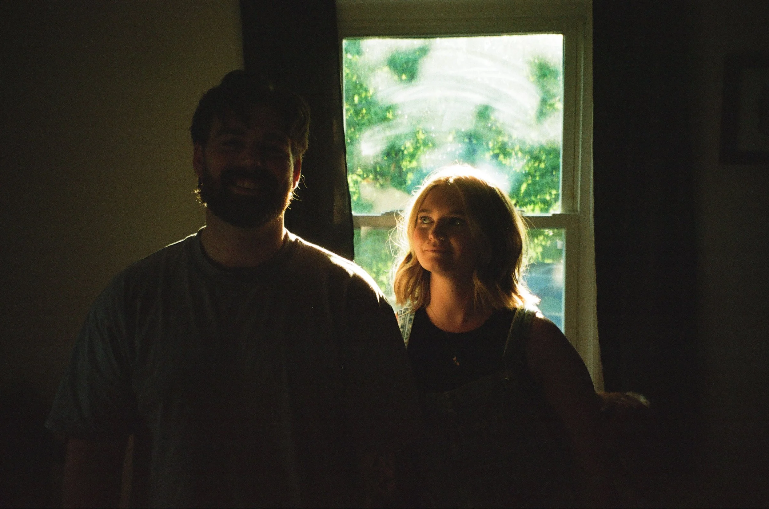 A man and woman are standing inside near a window with sunlight streaming in behind them. Their faces are silhouetted, with the woman's face partially illuminated and showing a gentle expression.