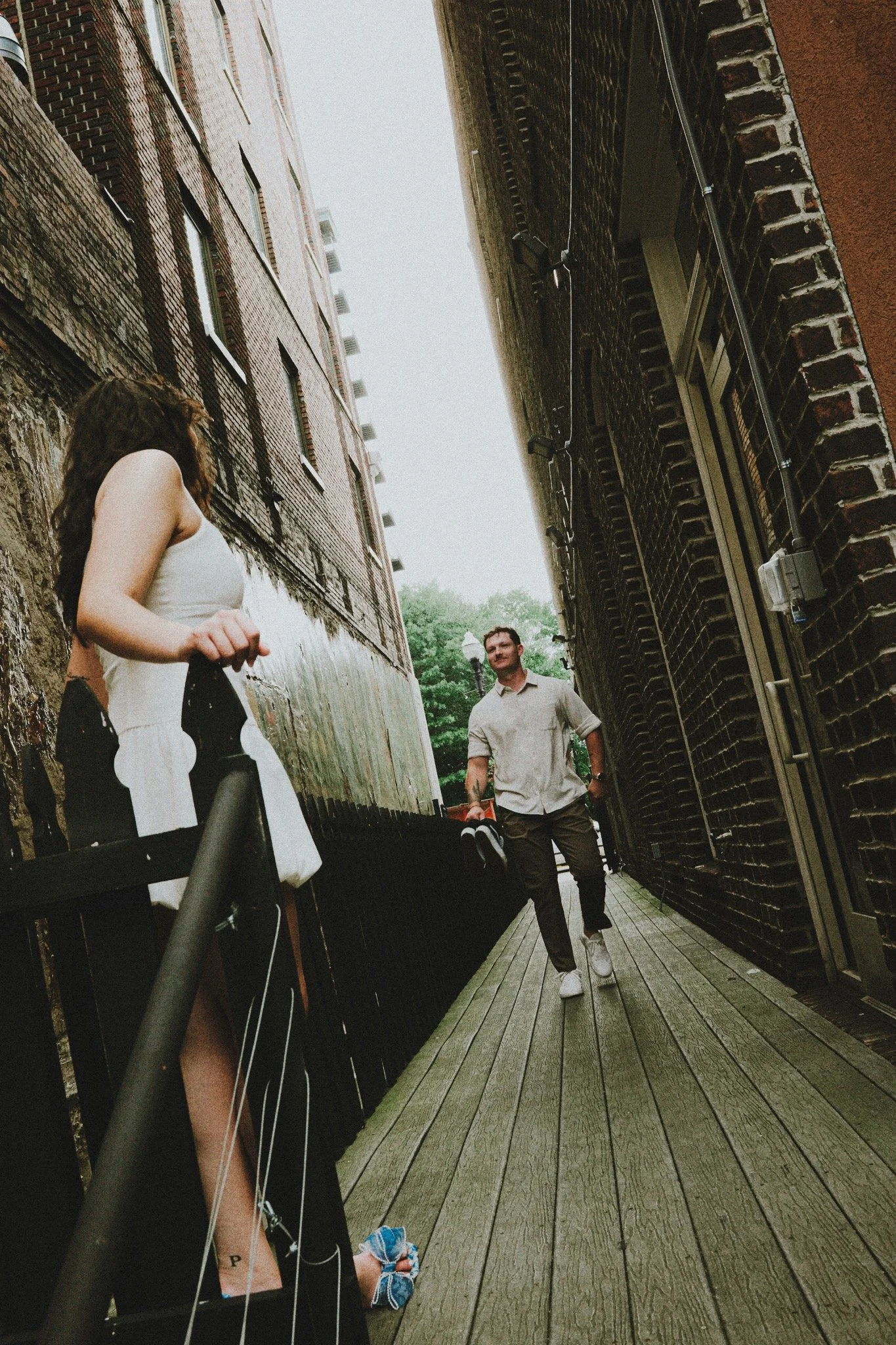 A woman standing on a balcony railing, holding a drink, looking towards a man walking on a wooden pathway between brick and stone buildings.