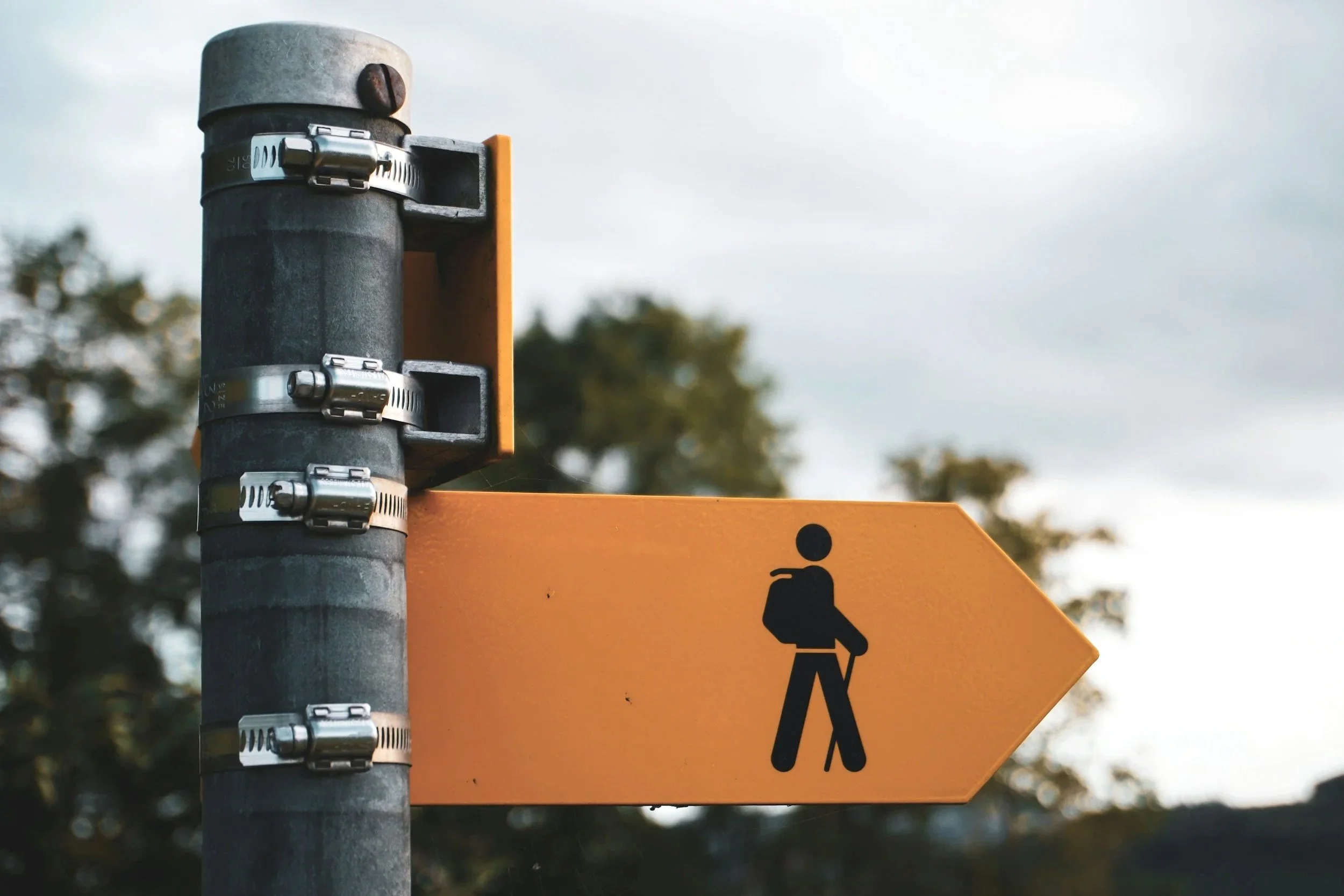 A yellow directional sign with a black walking person symbol, mounted on a dark metal pole with clamps.