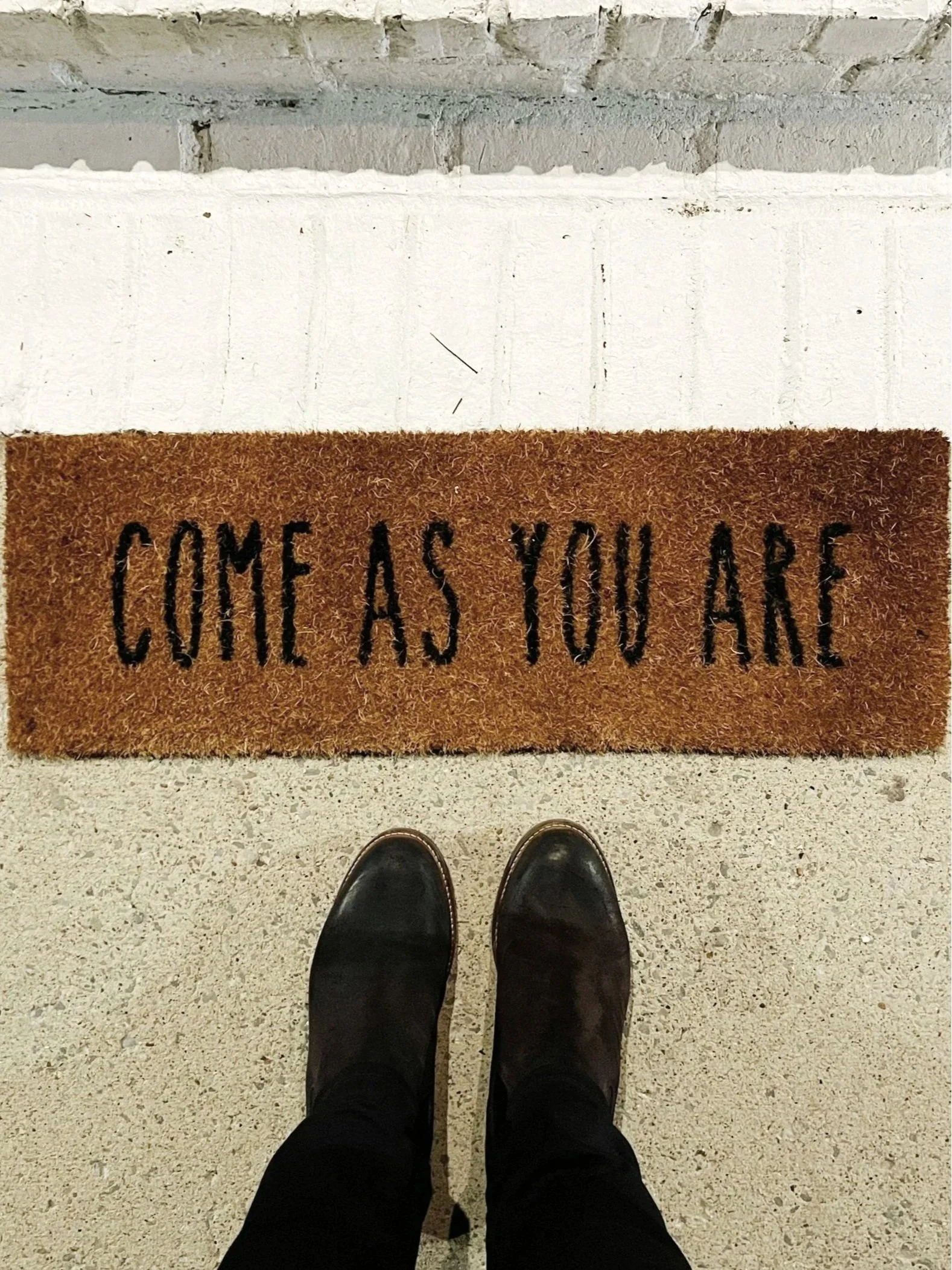 A person's feet in black shoes standing on a light-colored floor in front of a brown doormat with black text that reads "COME AS YOU ARE". The doormat is placed on a concrete or cement surface.