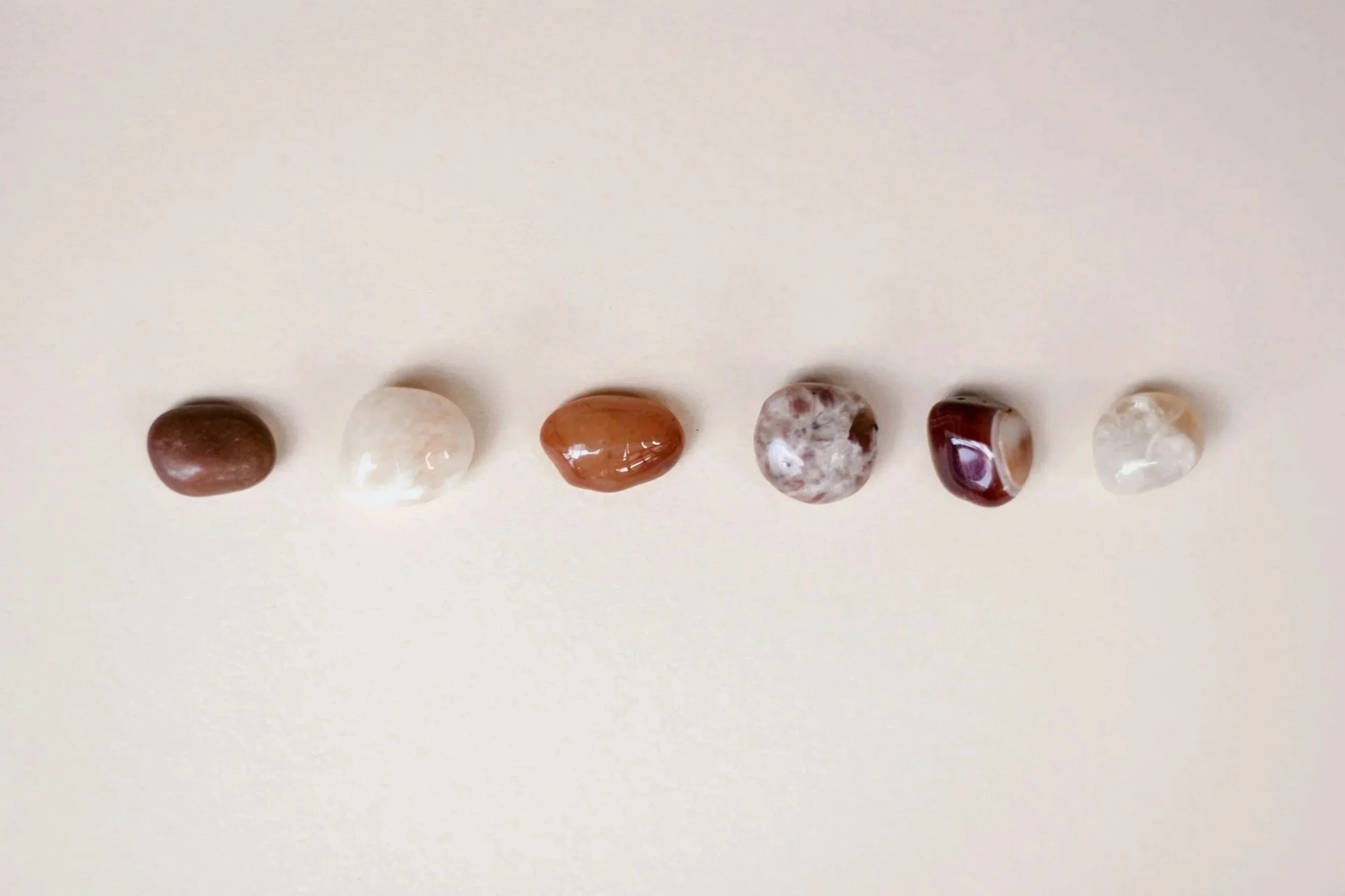 Seven polished tumbled stones of various colors and patterns arranged in a straight line on a white background.