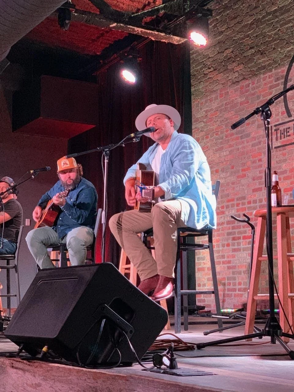 Two musicians performing on stage with guitars, one wearing a white hat and light blue shirt, and the other wearing a baseball cap and denim jacket, in a club with brick wall background.