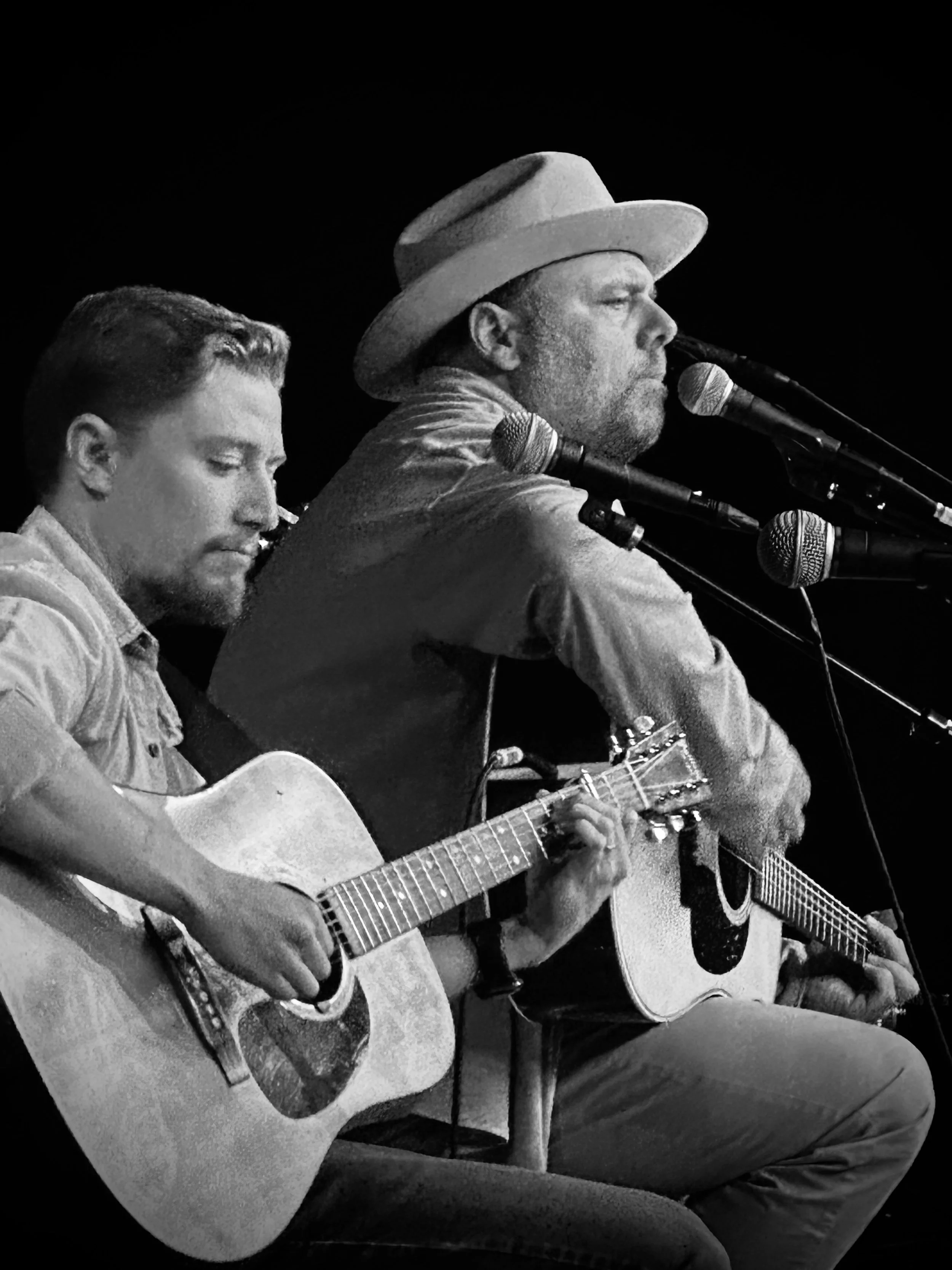 Neil Medley and Timothy Baker perform with acoustic guitars and microphones, one wearing a hat and singing passionately, the other with a focused expression, on a dark stage.