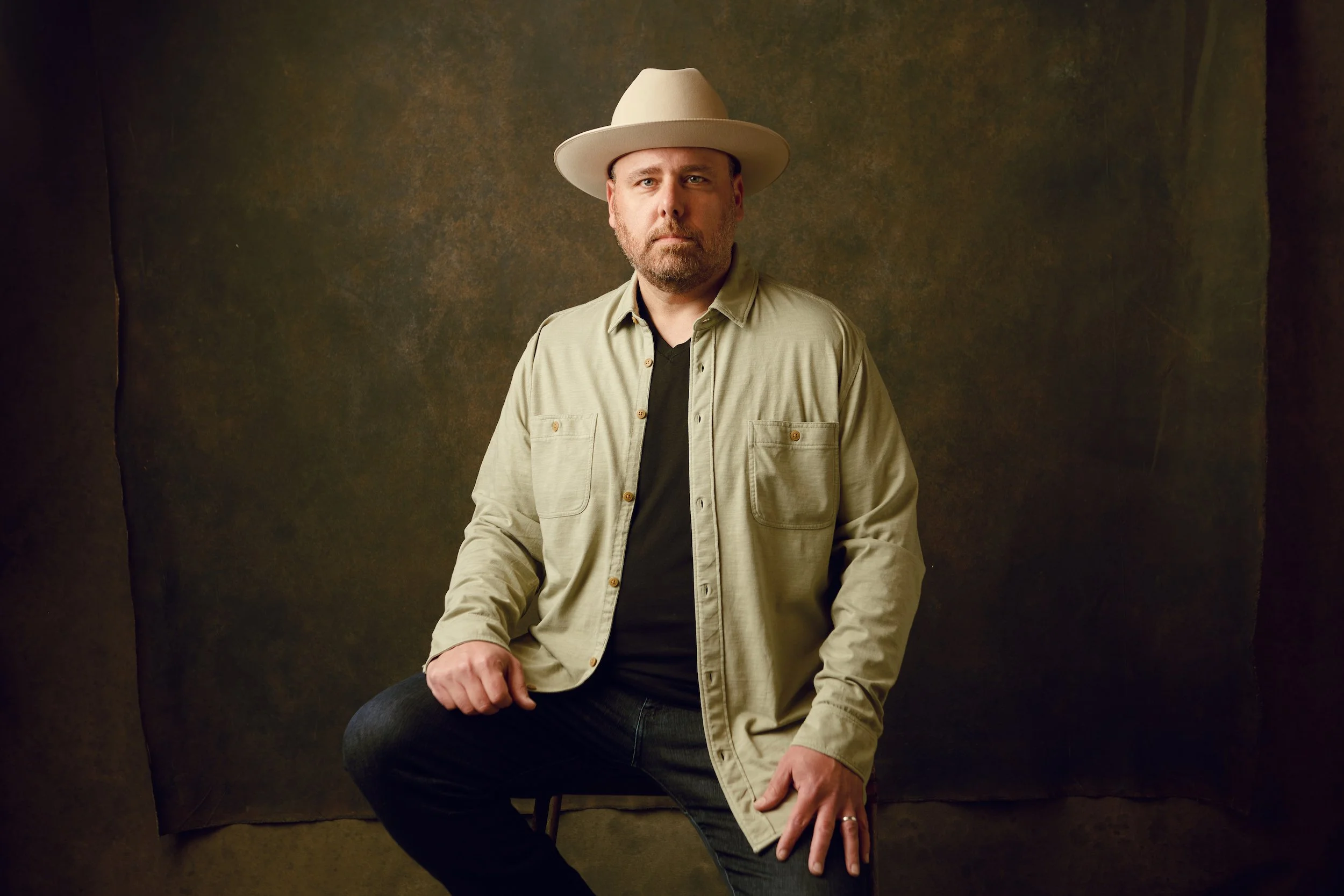 A man with a beard wearing a beige hat, a beige button-up shirt, and a black t-shirt, sitting against a dark, textured background.