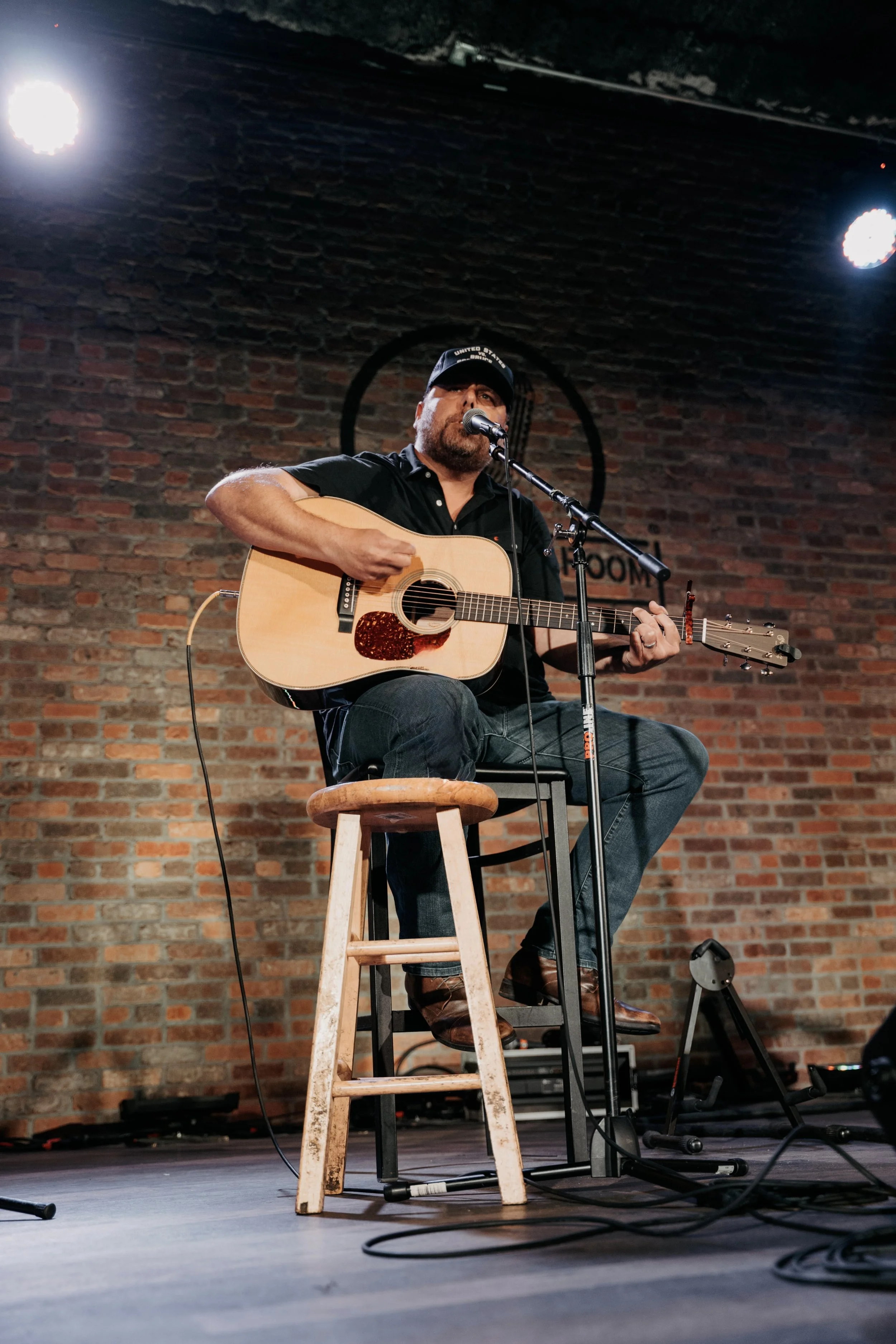 Neil Medley sitting on a stool playing an acoustic guitar and singing into a microphone on a stage with a brick wall background.