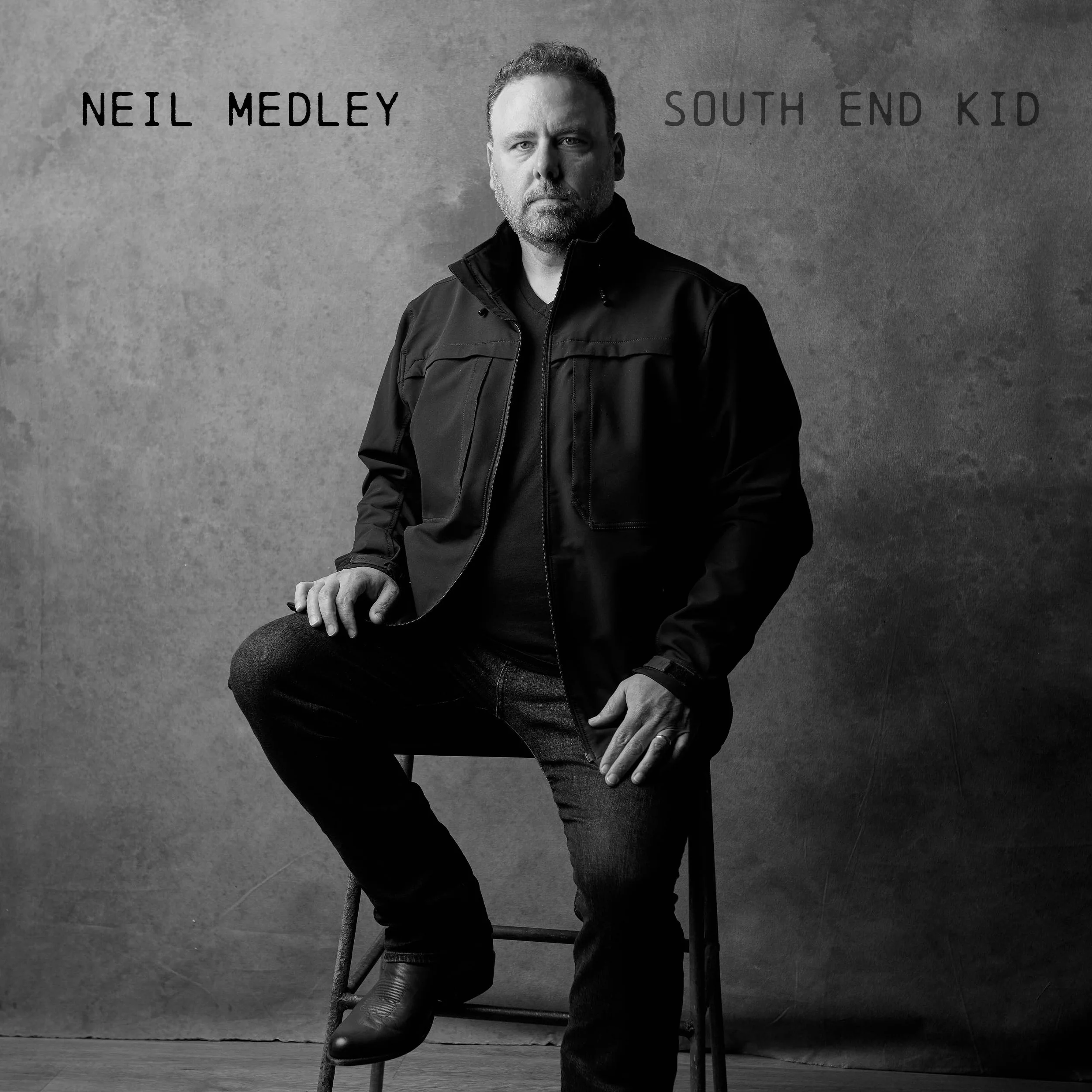 Black and white photo of Neil Medley sitting on a stool against a textured wall, wearing a dark jacket and jeans, with text indicating the name 'Neil Medley' and 'South End Kid'.