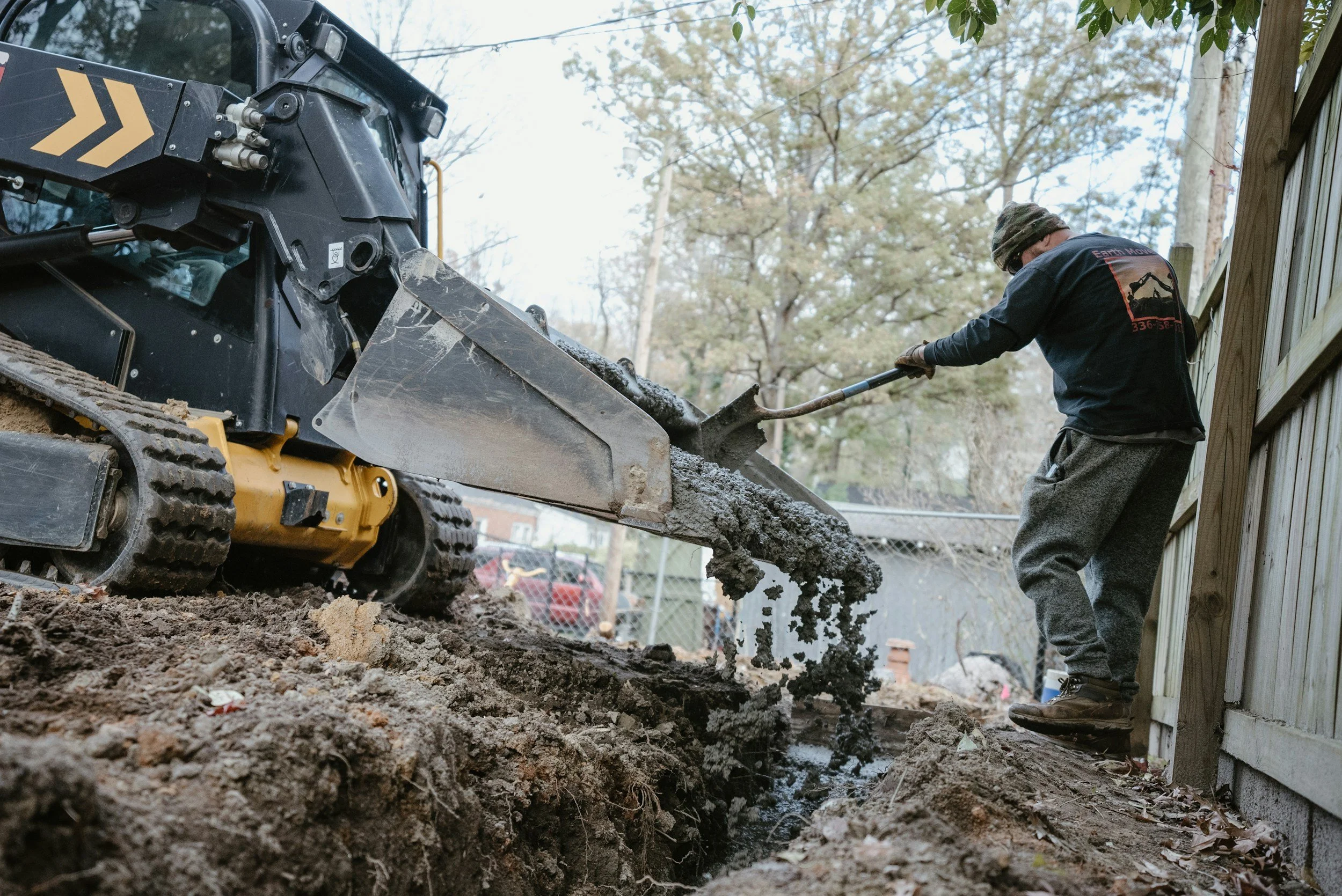 A person operating skid-steer in a backyard fillinging a footer with concrete, with their foot on the ground and a fence in the background.