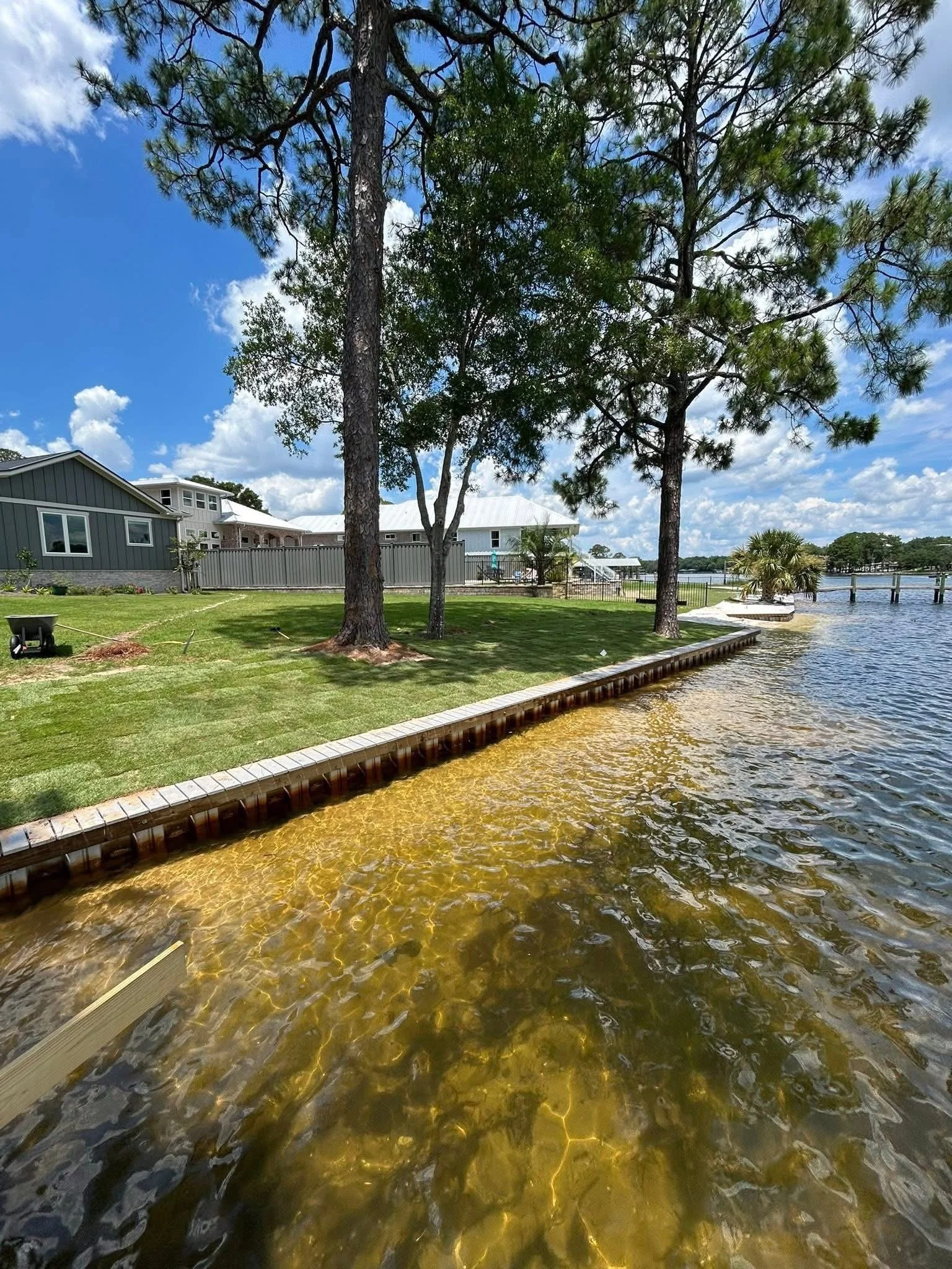 Bayside backyard with tall trees, freshly laid green sod, a sea-wall, and waterfront view, under a partly cloudy sky.