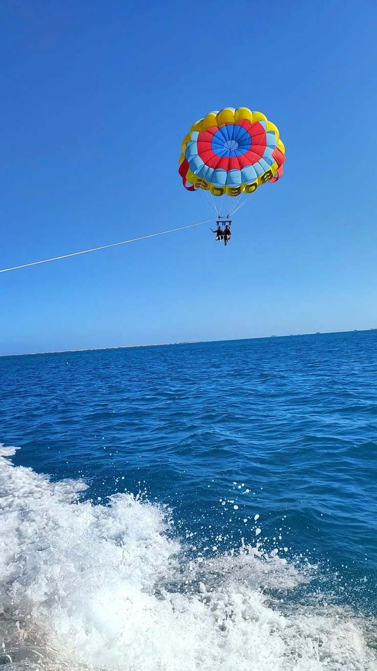 Parachute coloré orange, jaune, rouge, et bleu dans un ciel bleu au-dessus de la mer. Deux personnes assises dans une nacelle de parachute. Vague d'eau en bas. C'est à l'île Maurice.