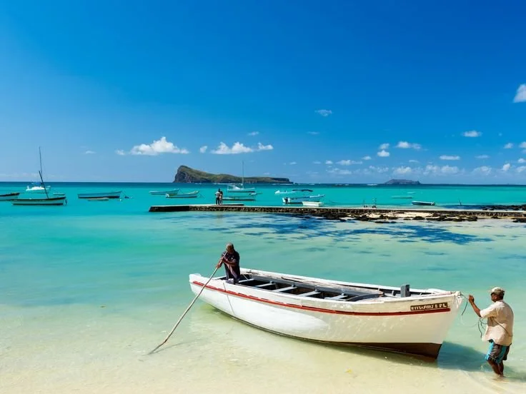 Deux personnes près d'un bateau blanc à la plage avec d'autres bateaux au large et une île en arrière-plan sous un ciel bleu. C'est à l'île Maurice.