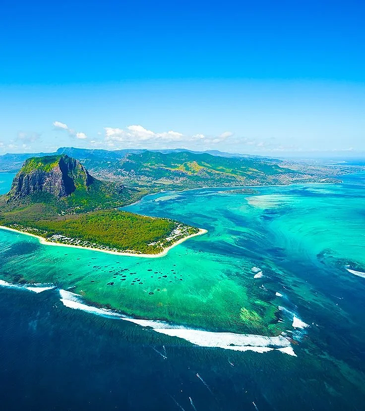 Vue aérienne d'une île tropicale avec une forêt dense, une plage de sable blanc, et des eaux turquoise entourant l'île, avec des montagnes au loin et un ciel bleu clair. C'est à l'île Maurice.