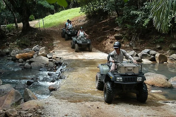 Quatre personnes faisant de la conduite tout-terrain avec des quads à travers une rivière en forêt. C'est à l'île Maurice.