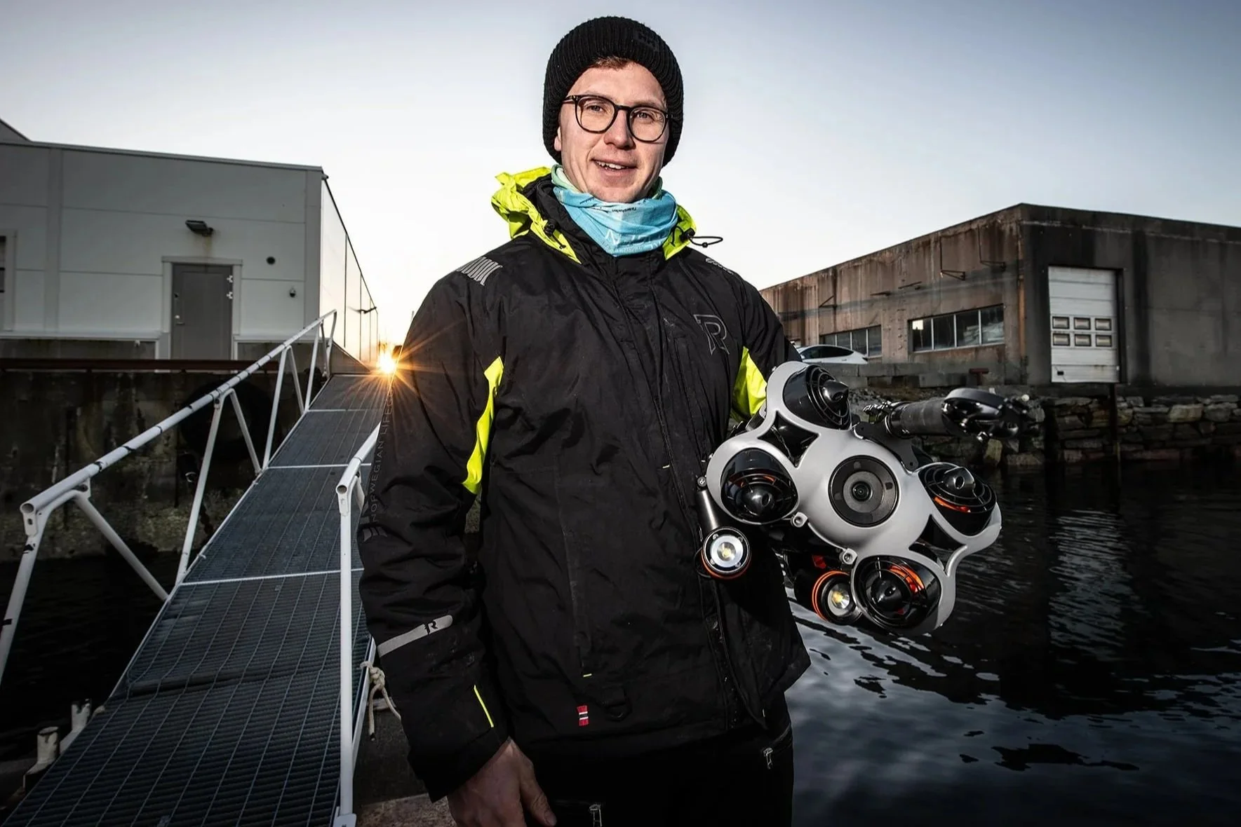 Martin Helseth står i flytejakken sin ved vannet, holder en moderne ROV med gripeklo i armene, med bygninger på Valderøya i bakgrunnen. Soloppgang