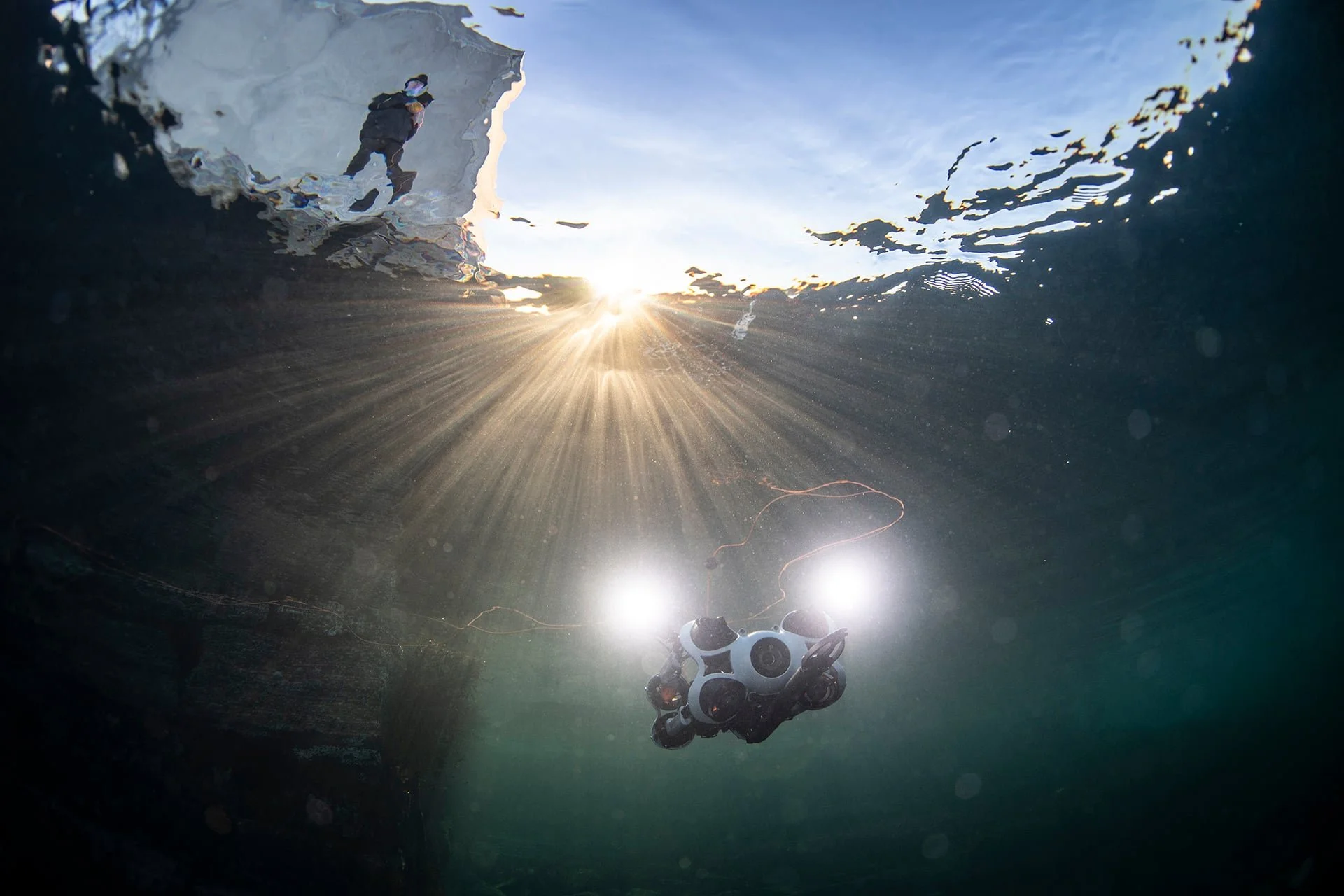 Undervannsbilde av en robot (ROV) med lys, der en person står på kaien i bakgrunnen. Sollyset trenger gjennom. Sikten er god