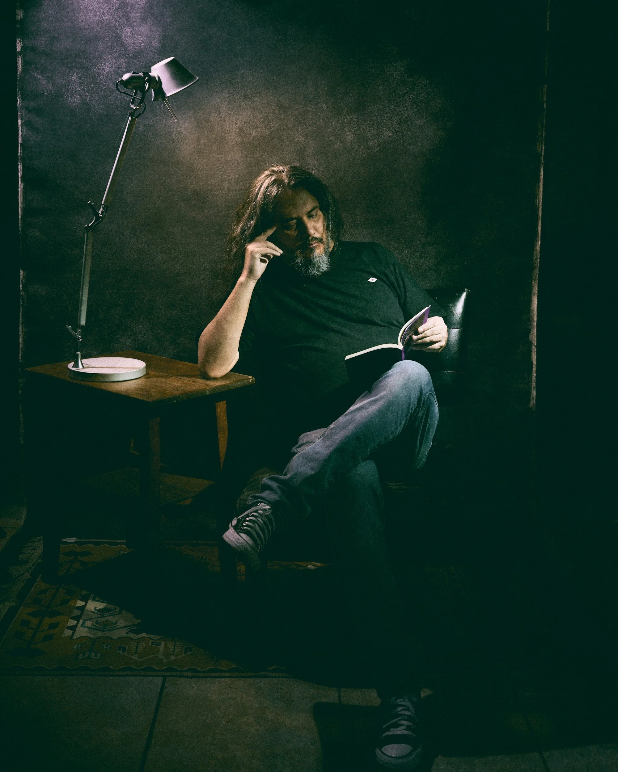 A man with long hair and a beard sitting in a dimly lit room reading a book, resting his head on his hand near a small wooden table with a modern desk lamp.