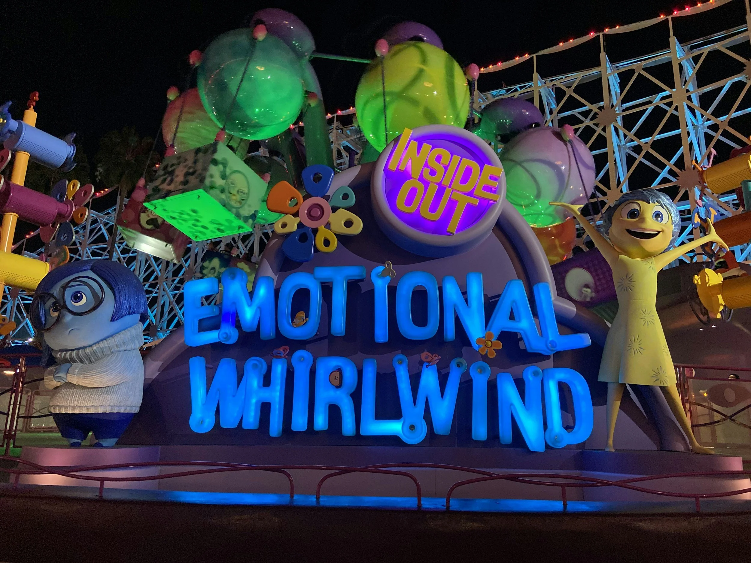 Colorful amusement park ride at night with large neon sign reading 'Emotional Whirlwind' and 'Inside Out' theme. Characters from the movie 'Inside Out' are nearby, including Joy with yellow dress smiling with arms outstretched and Sadness with glasses and a sweater looking thoughtful.