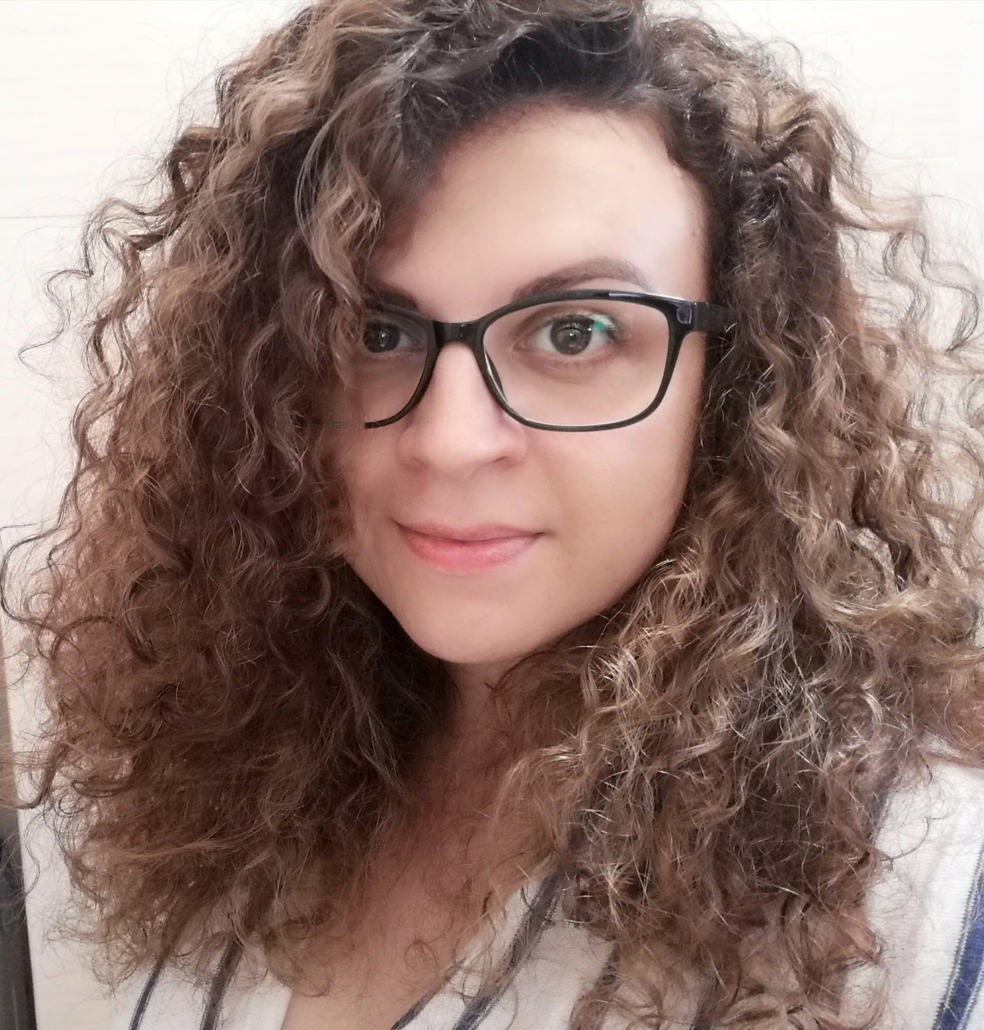 A woman with curly brown hair, wearing black glasses, smiling, in front of a light-colored wall.