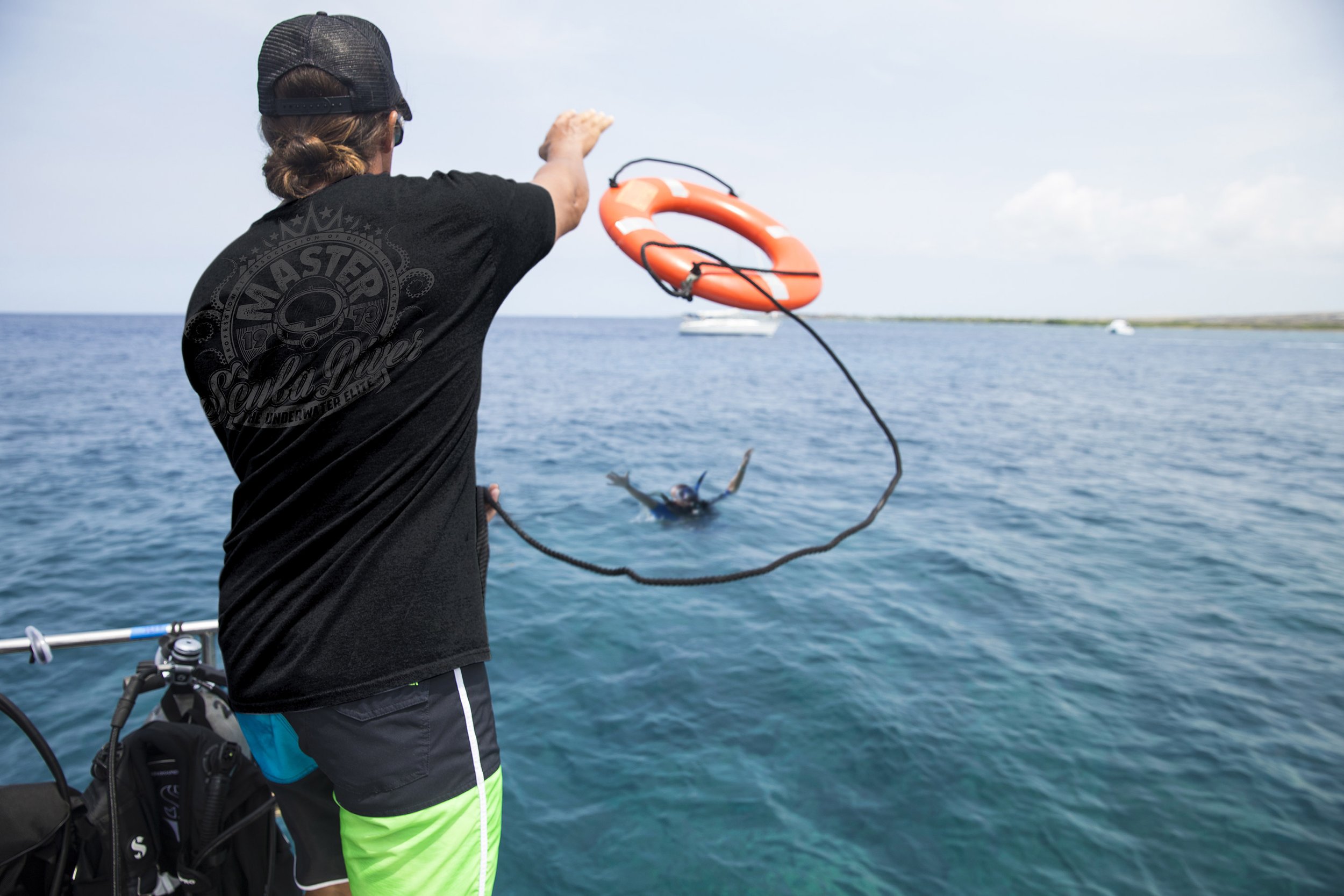 A person is fishing from a boat in a large body of water, holding a fishing line with a fish on the hook. The individual is wearing a black cap, black t-shirt, and bright green shorts. There is an orange lifesaver ring attached to the fishing line.