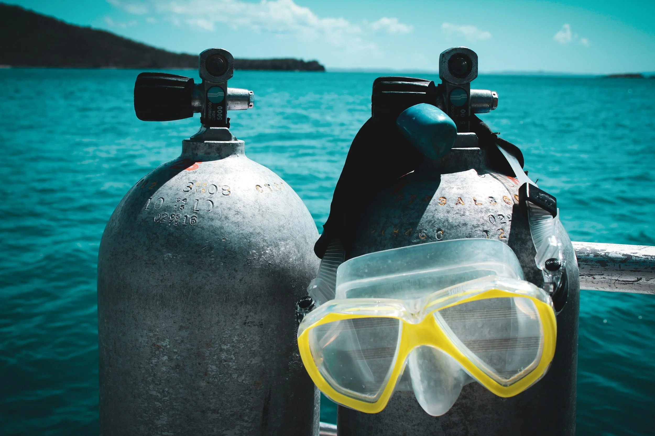 Two scuba tanks are positioned on the edge of a boat in front of turquoise ocean water with a distant shoreline and partially cloudy sky in the background. A pair of yellow scuba diving goggles and a snorkel are placed on the tank on the right.