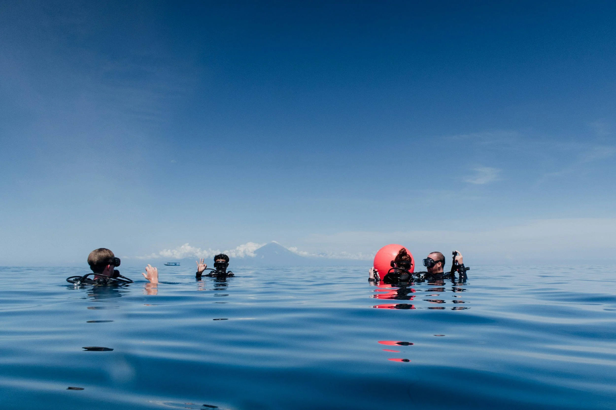 People scuba diving in the ocean, with a clear sky and mountain in the background, some of them waving and taking pictures.