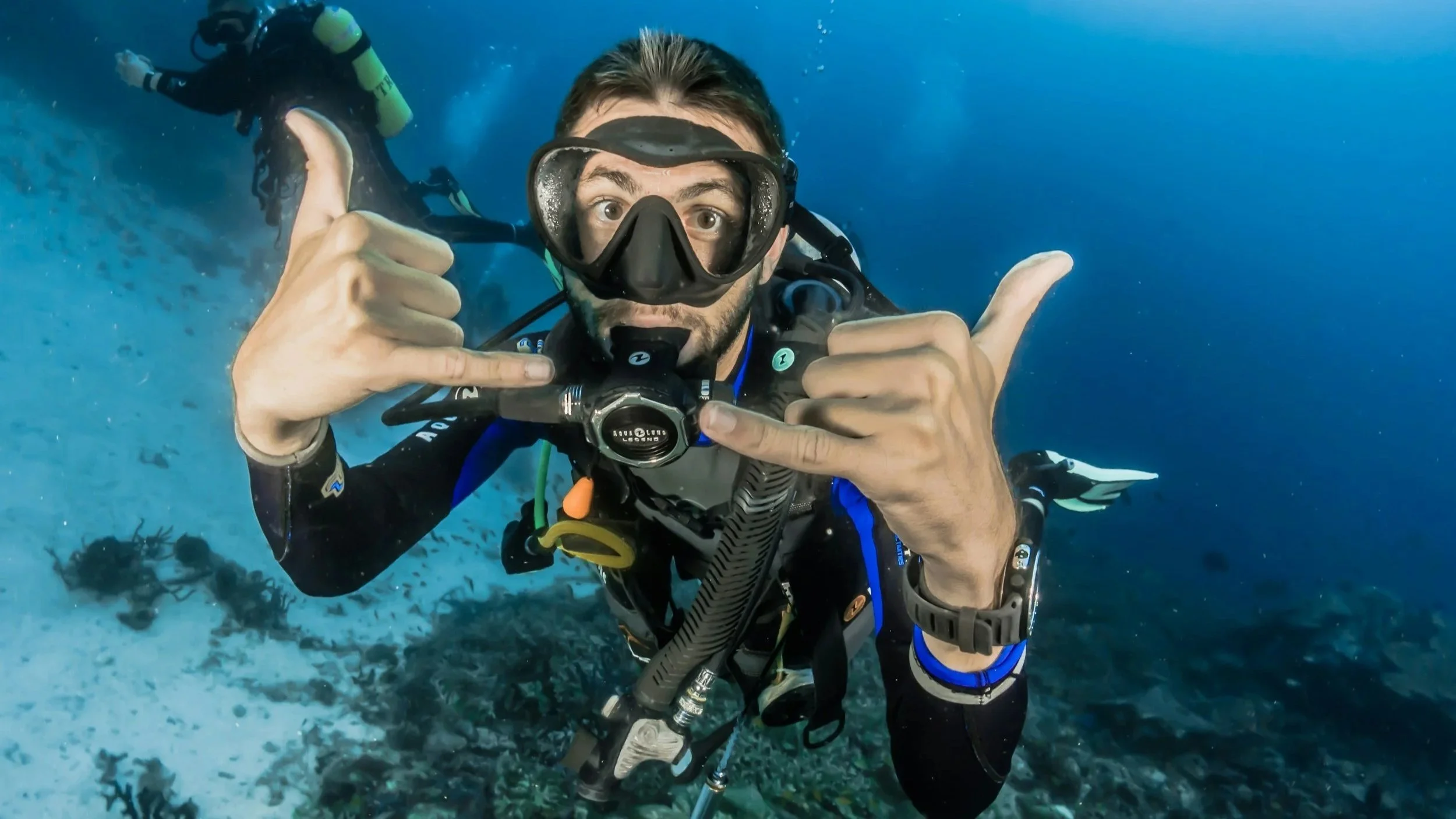 A man wearing scuba diving gear underwater, giving a thumbs-up and making the 'hang loose' gesture with both hands, with another diver visible in the background.
